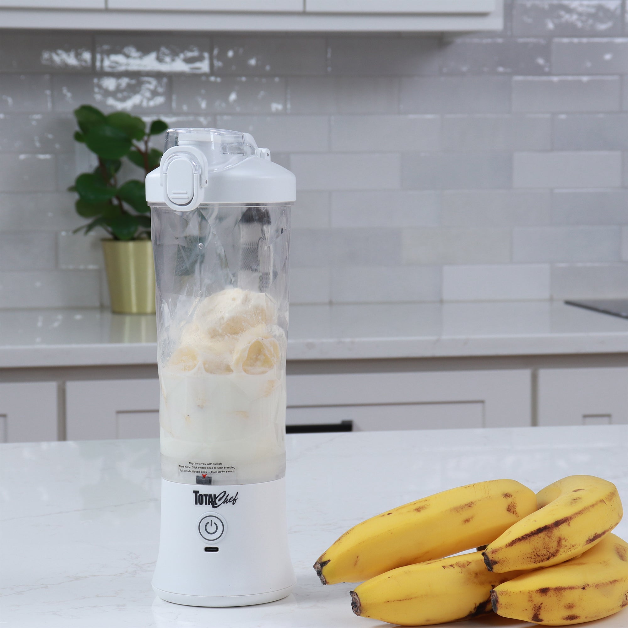 Lifestyle image of the Total Chef USB chargeable blender filled with milk and slices of banana on a white marble counter with a bunch of bananas beside it and a light gray tiled backsplash behind.
