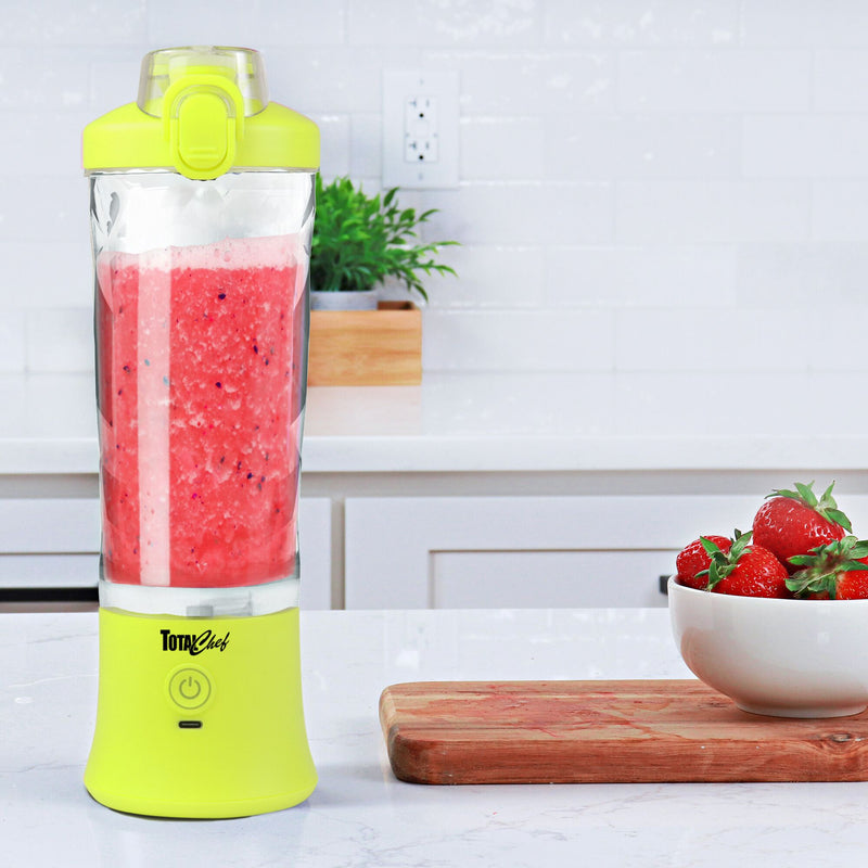 Lifestyle image of the Total Chef portable rechargeable blender filled with bright pink smoothie in a white kitchen with a wooden cutting board and a white bowl of strawberries beside it.