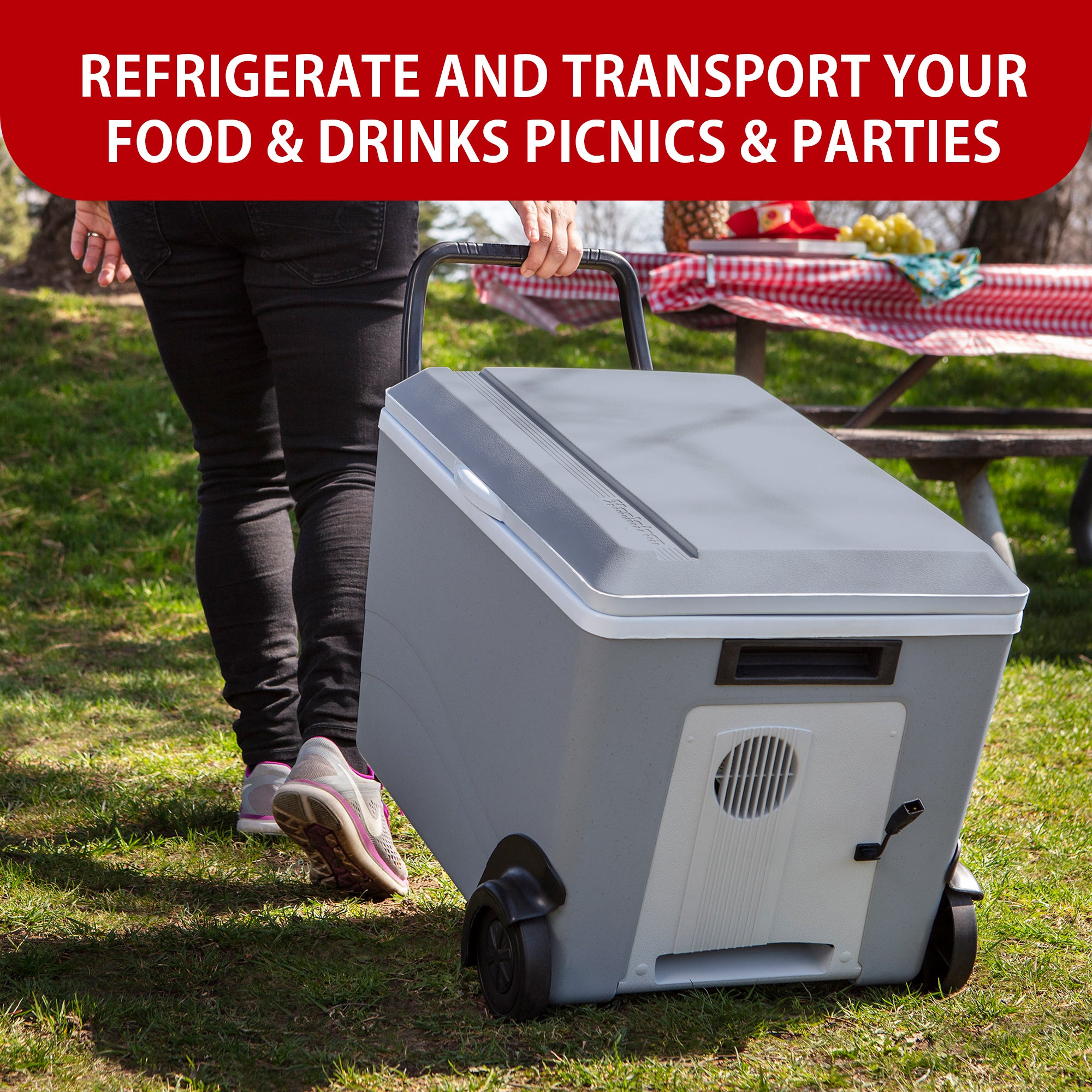 A person wearing black pants and gray and pink running shoes is towing the Koolatron rolling 12V cooler/warmer on grass towards a picnic table with a red and white checked tablecloth. Text above reads, "REFRIGERATE AND TRANSPORT FOOD AND DRINKS TO PICNICS AND PARTIES"