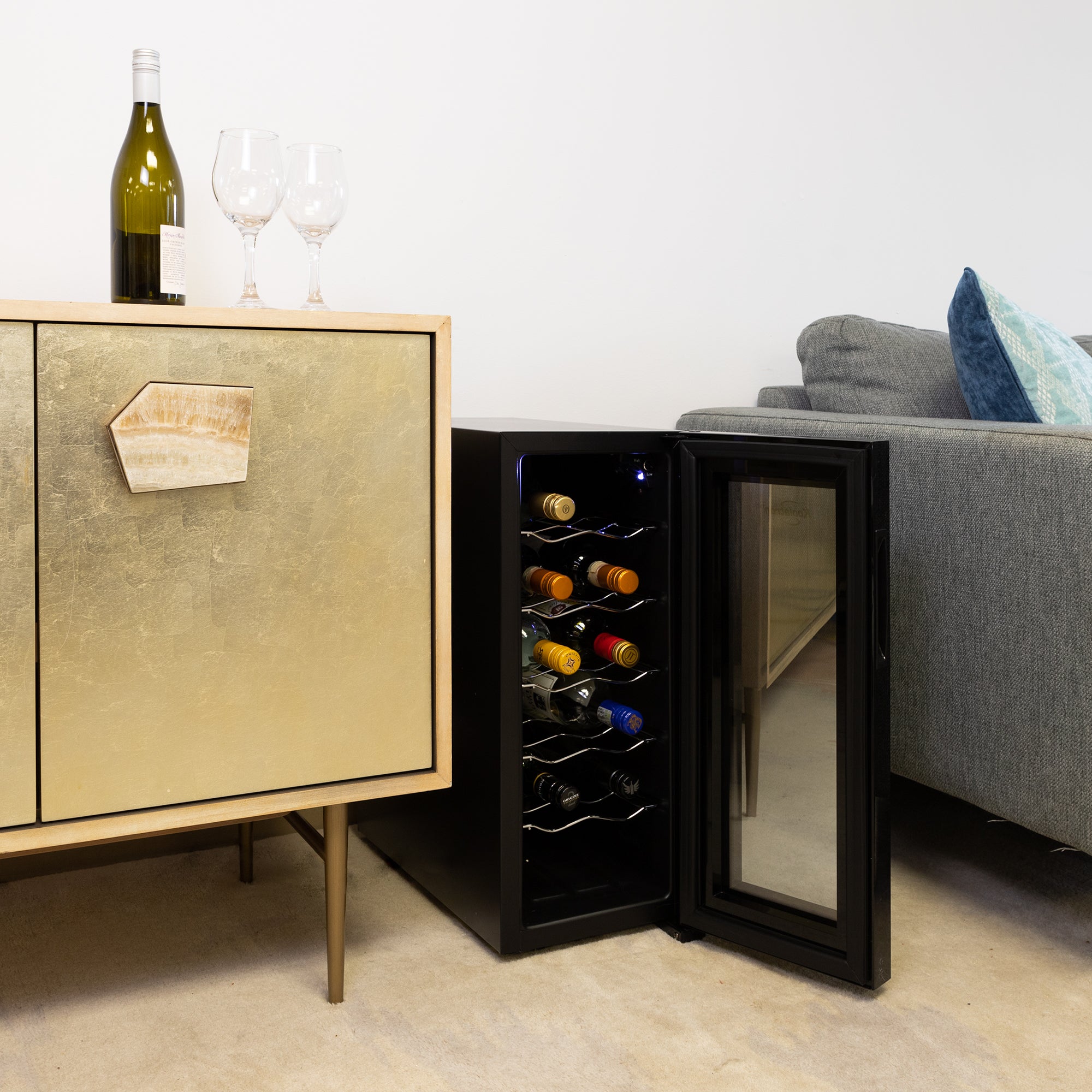 Koolatron 12 bottle wine cooler, open and filled with bottles of wine, set up on the floor between a gray sofa and a gold-coloured sideboard with a bottle of wine and one glass beside it