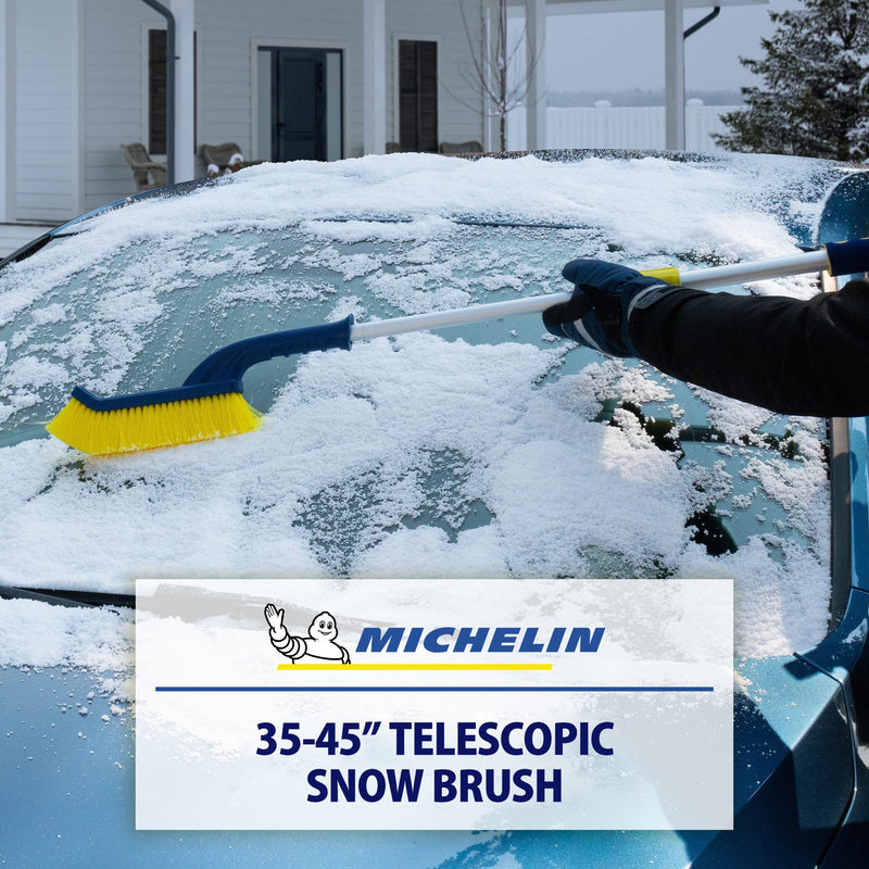 Lifestyle image of a gloved hand using the snow brush, extended, to remove snow from the windshield of a dark coloured car. Transparent white overlay at the bottom shows the Michelin logo above text reading "35-45" telescopic snow brush"