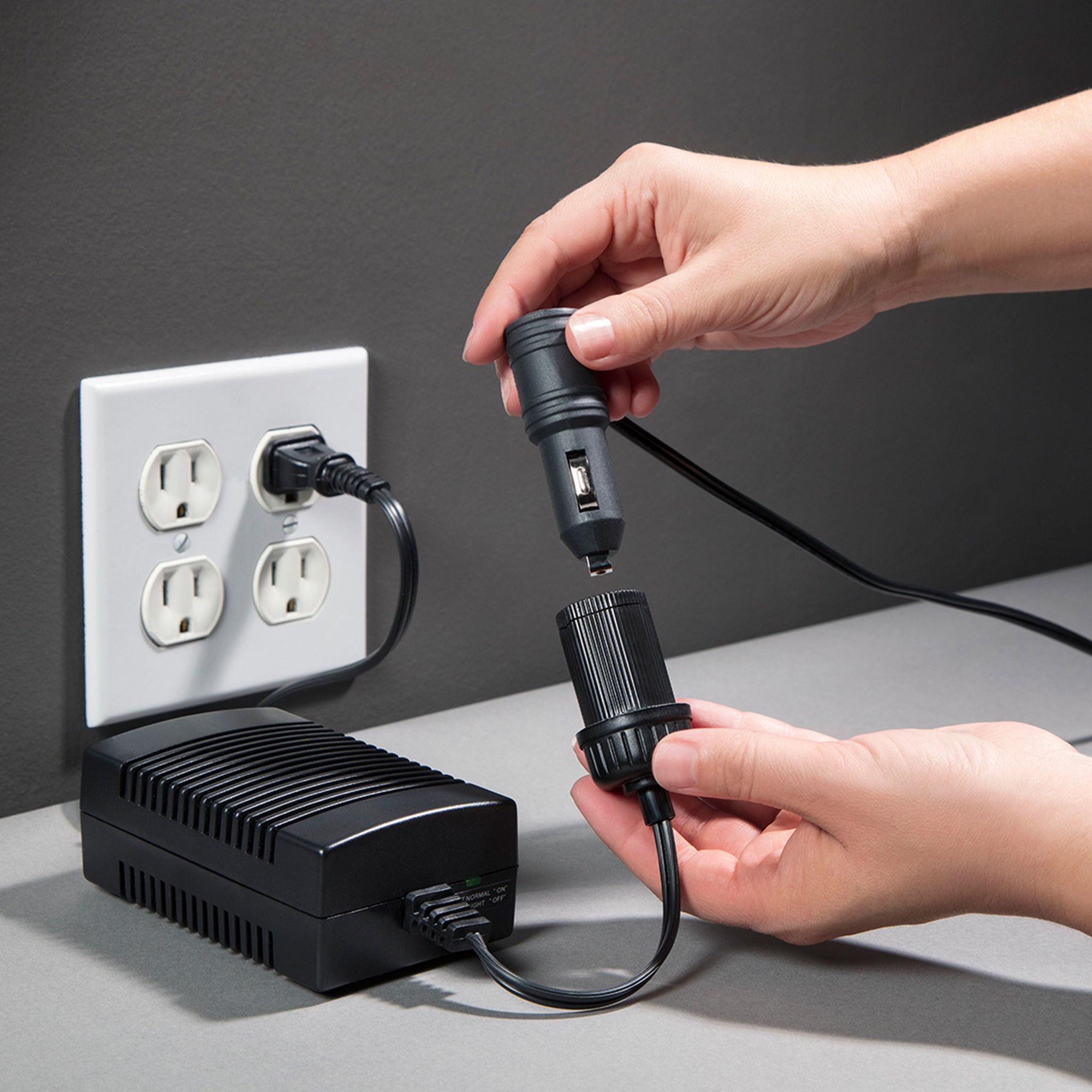 Lifestyle image of the AC to DC power adapter plugged into a white indoor AC receptacle on a dark gray wall with a person’s hands inserting a 12V plug into the DC adapter end