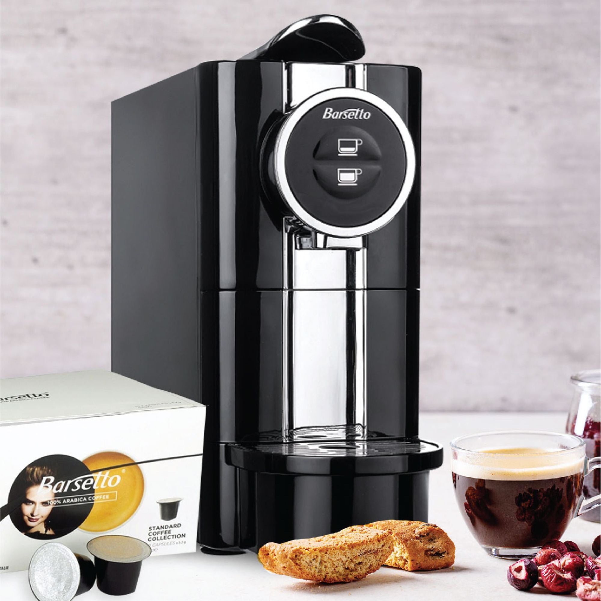 Lifestyle image of Barsetto one-touch espresso machine on a white countertop with a light gray backsplash behind and a box of espresso pods, pieces of biscotto, and a shot of espresso in a clear glass mug arranged around it