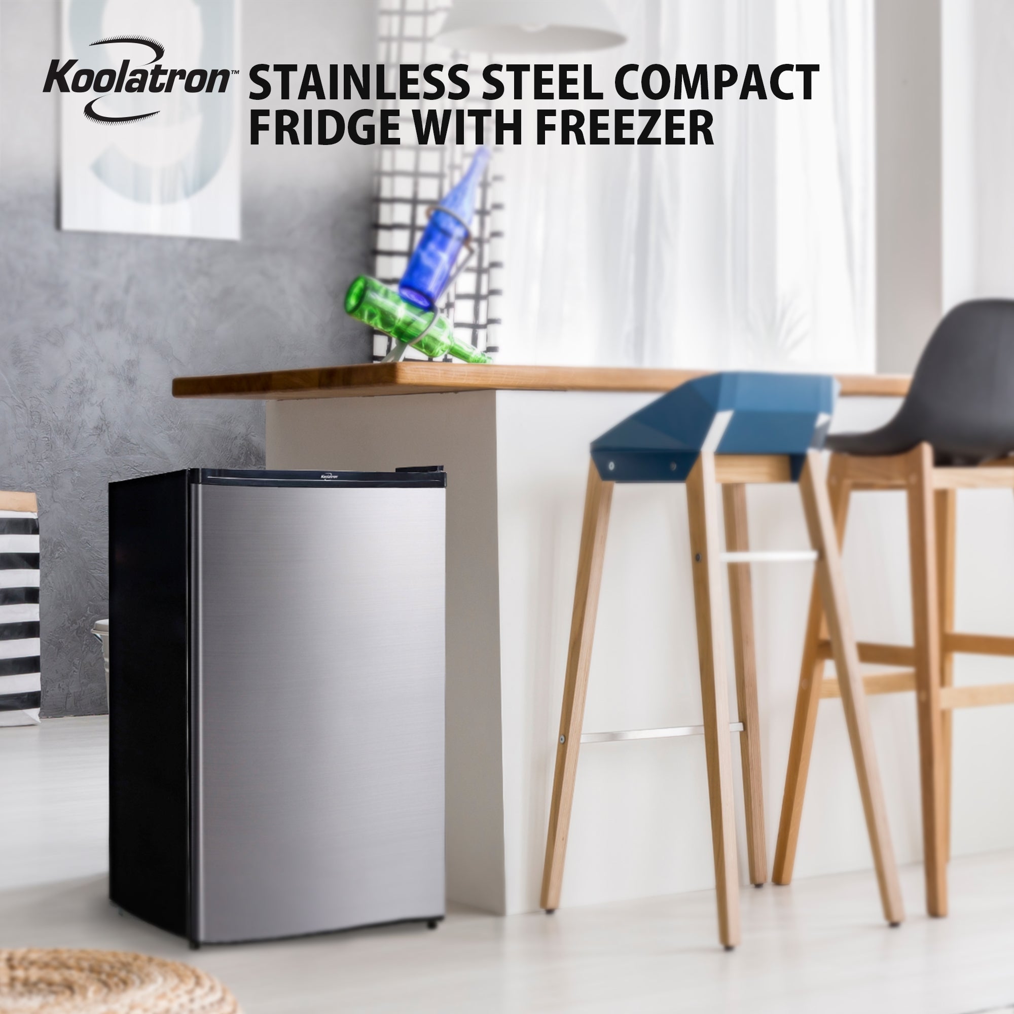 Lifestyle image of black and stainless steel compact fridge with freezer beside a white breakfast bar with wooden countertop and two wood and plastic bar stools. Text above reads "Koolatron stainless steel compact fridge with freezer"