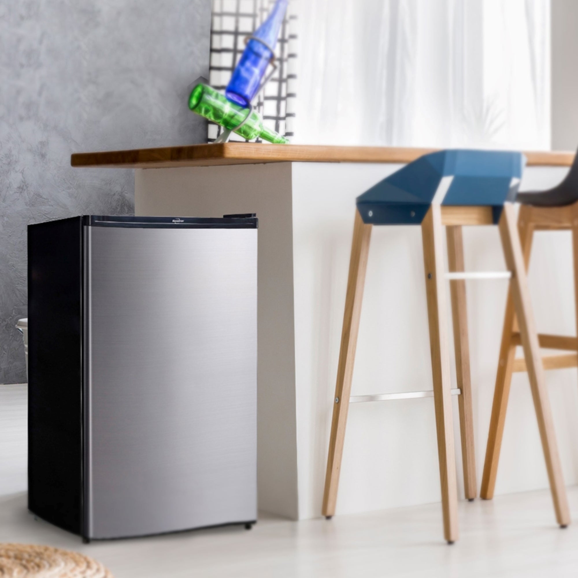 Lifestyle image of black and stainless steel compact fridge with freezer beside a white breakfast bar with wooden countertop and two wood and plastic bar stools. Text above reads "Koolatron stainless steel compact fridge with freezer"