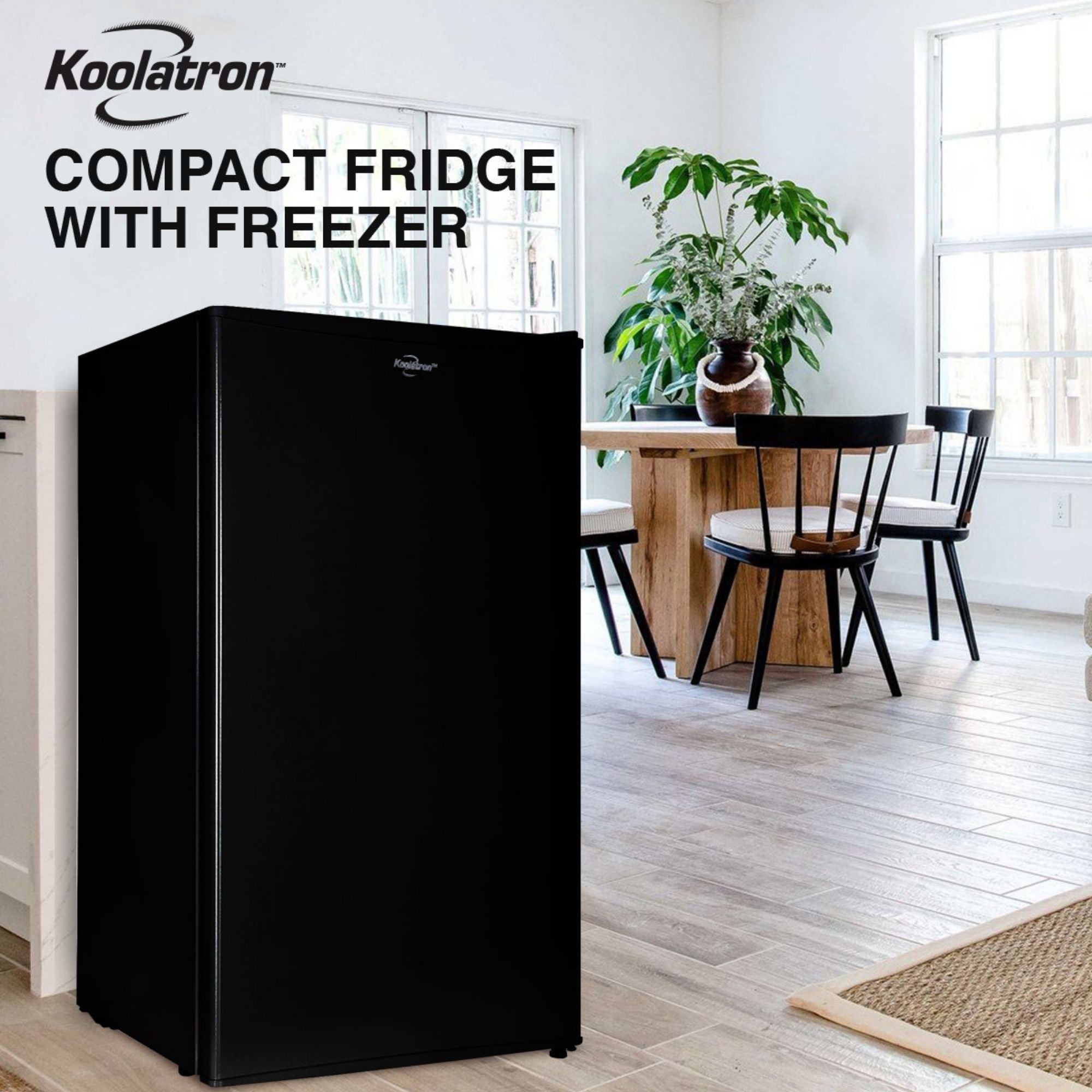 Lifestyle image of black compact fridge with freezer on a light-colored plank floor with a wooden table and chairs, two large windows, and a large potted plant in the background. Text above reads "Koolatron compact fridge with freezer"