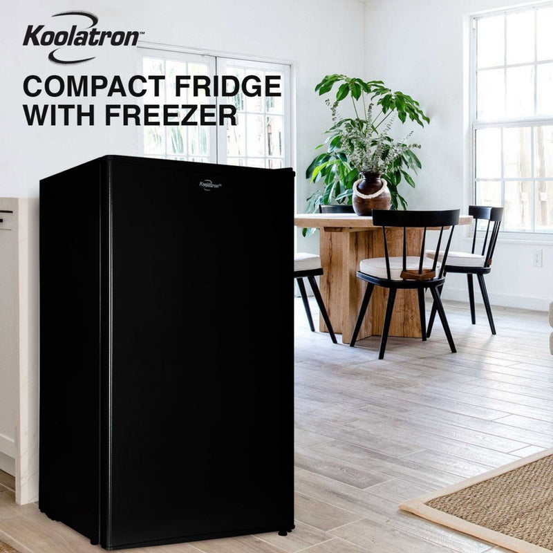 Lifestyle image of black compact fridge with freezer on a light-colored plank floor with a wooden table and chairs, two large windows, and a large potted plant in the background. Text above reads "Koolatron compact fridge with freezer"