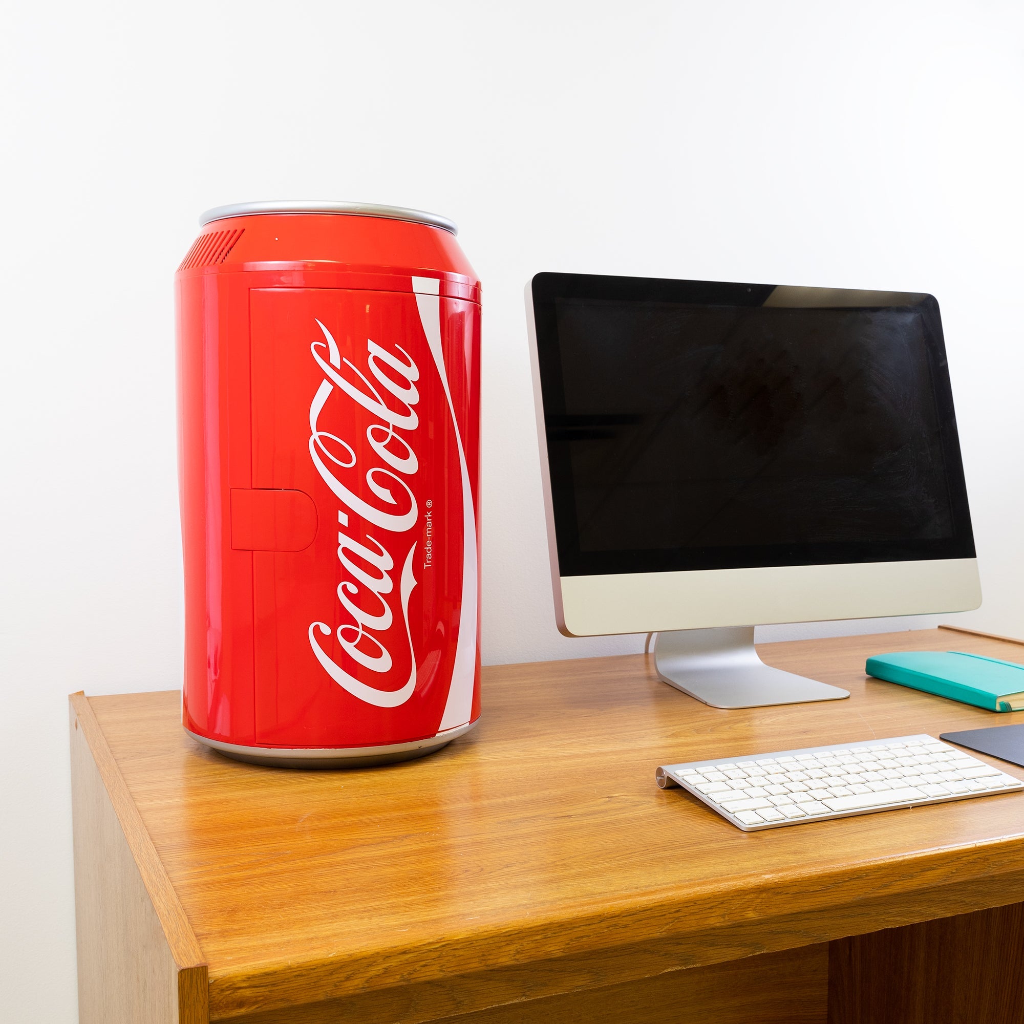 Lifestyle image of Coca-Cola can-shaped 12 can mini fridge, closed, on a wooden desktop with a computer monitor and keyboard beside and a white wall behind