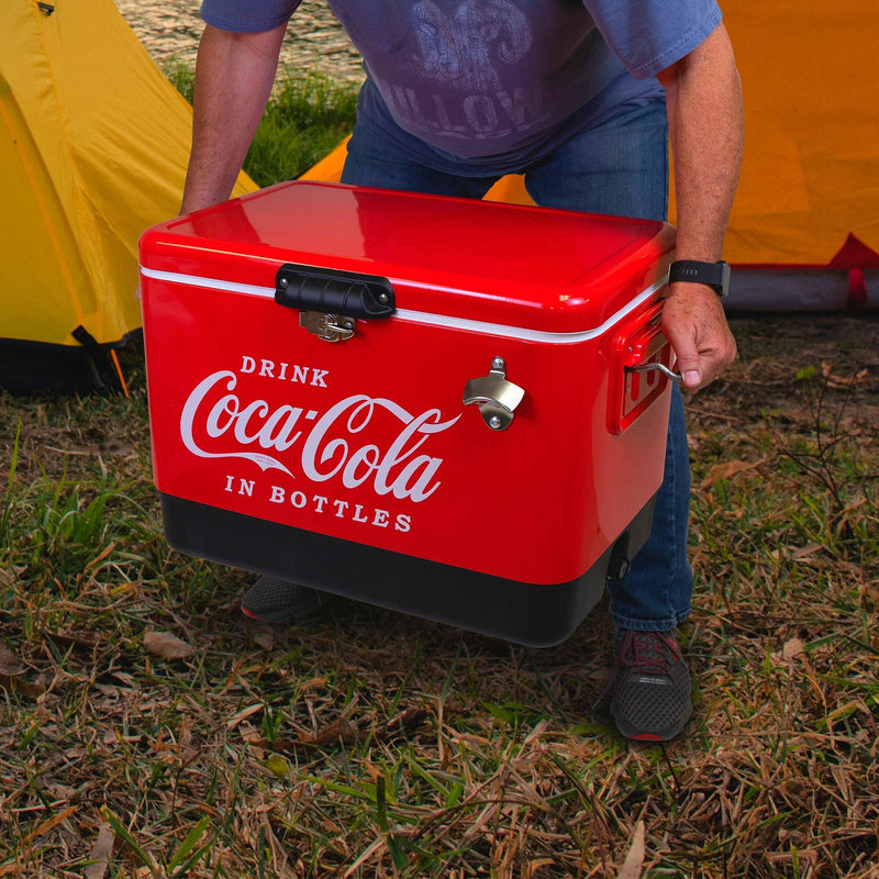 Lifestyle image of a person wearing jeans and a blue t-shirt lifting the Coca-Cola 54 quart ice chest with two yellow dome tents visible in the background
