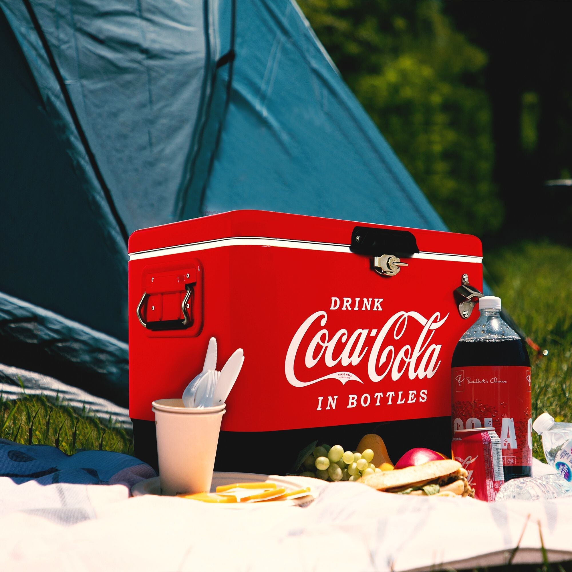 Lifestyle image of Coca-Cola 54 qt ice chest on a picnic blanket on the grass with fresh fruit and a bottle and can of Coke in front and a teal dome tent behind