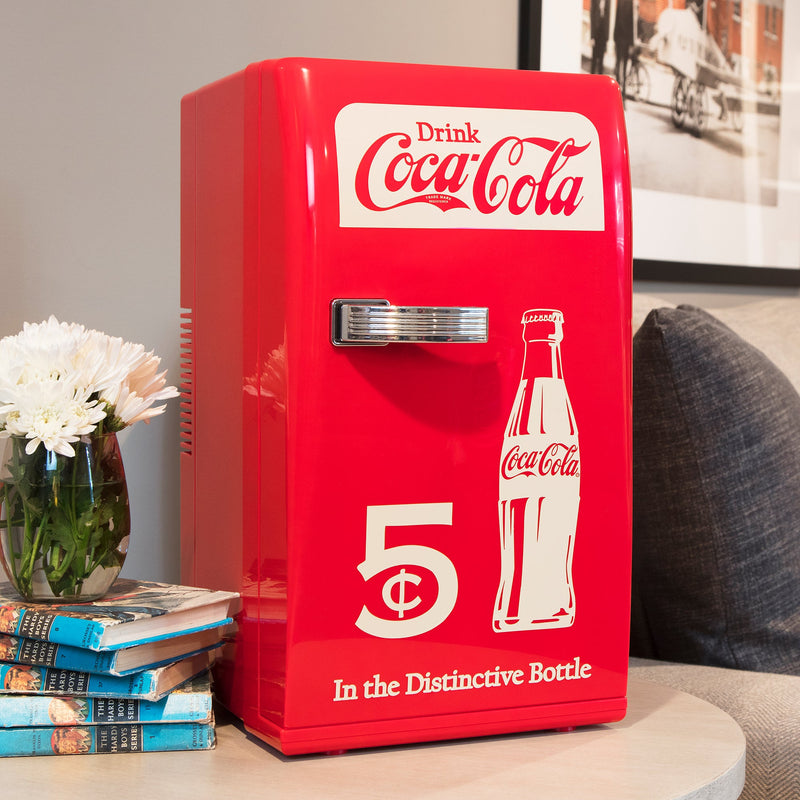 Lifestyle image of Coca-Cola retro 18 can mini fridge, closed, on a light-colored wooden side table with a sofa to the right and a stack of hardcover books and vase of white flowers to the left