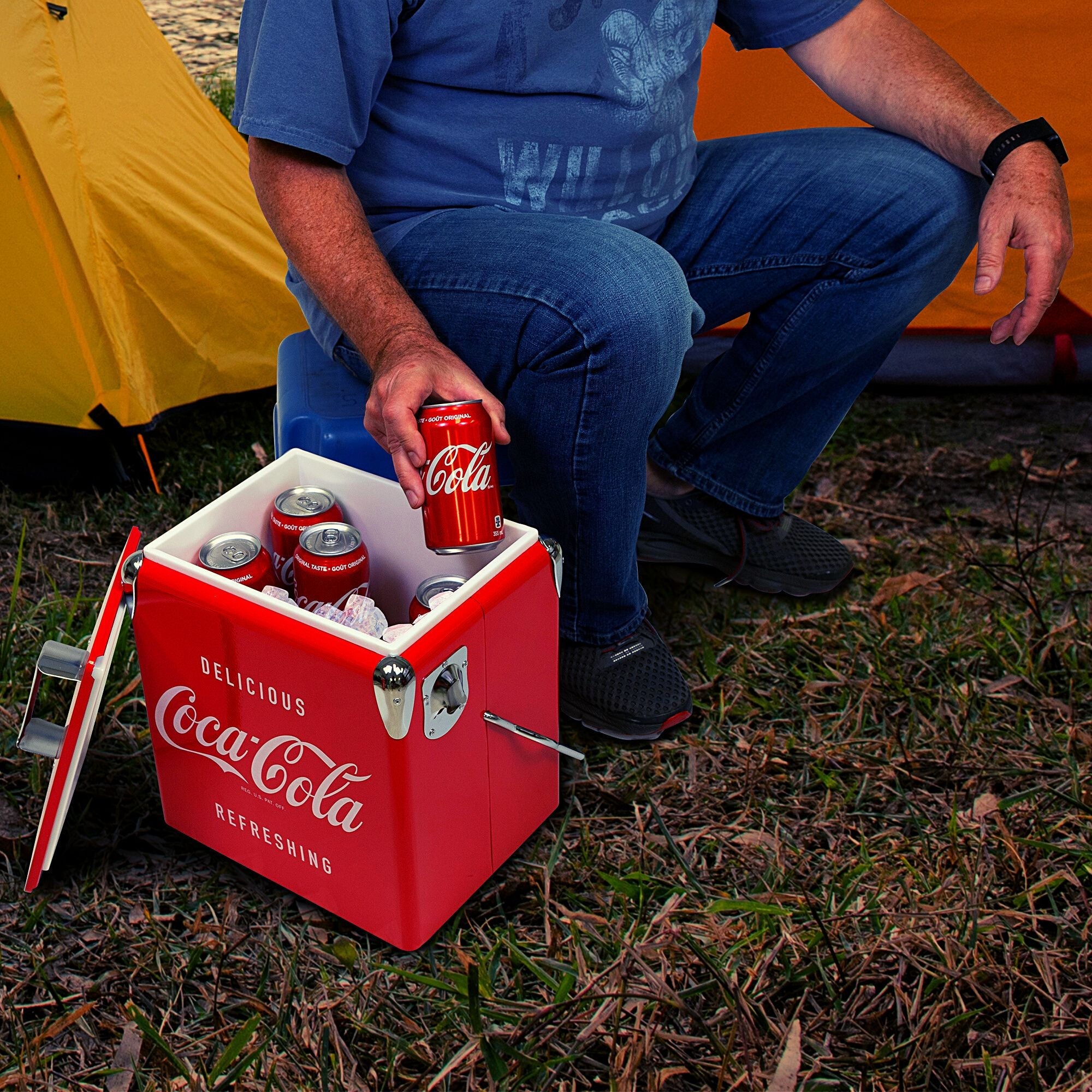Lifestyle image of a person wearing jeans and a blue t-shirt sitting in front of two yellow dome tents and lifting a can of Coke out of the open the Coca-Cola retro ice chest