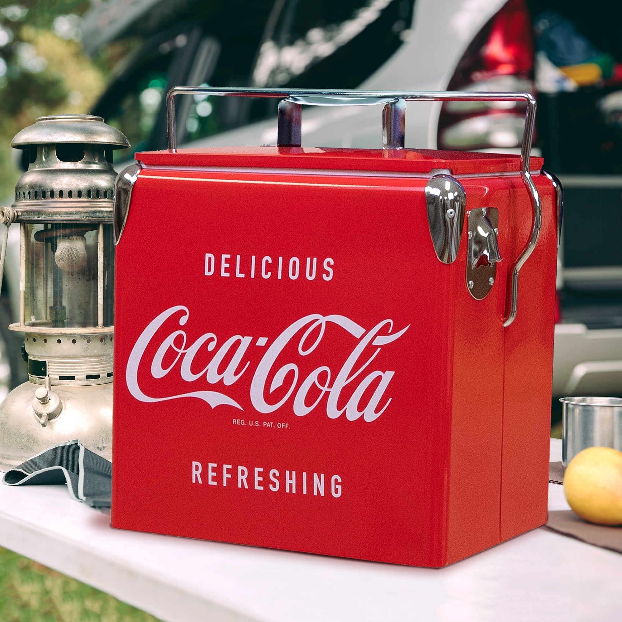 Lifestyle image of Coca-Cola retro ice chest with bottle opener on a white folding table with a metal lantern beside it and a vehicle in the background