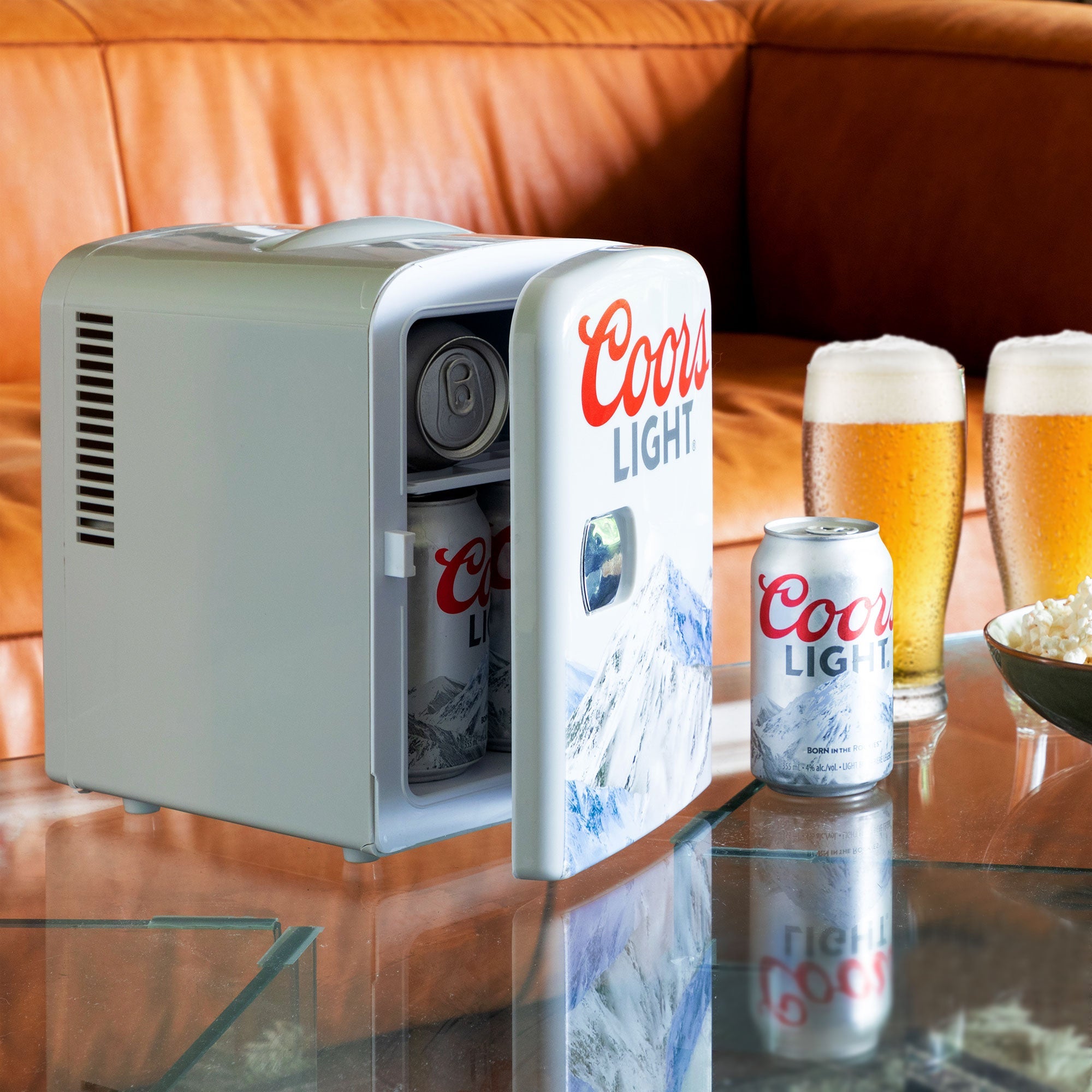 Lifestyle image of Coors Light portable mini fridge, partly open with cans visible inside, on a glass coffee table with a can and two glasses of Coors Light beside it and a tan leather sofa behind