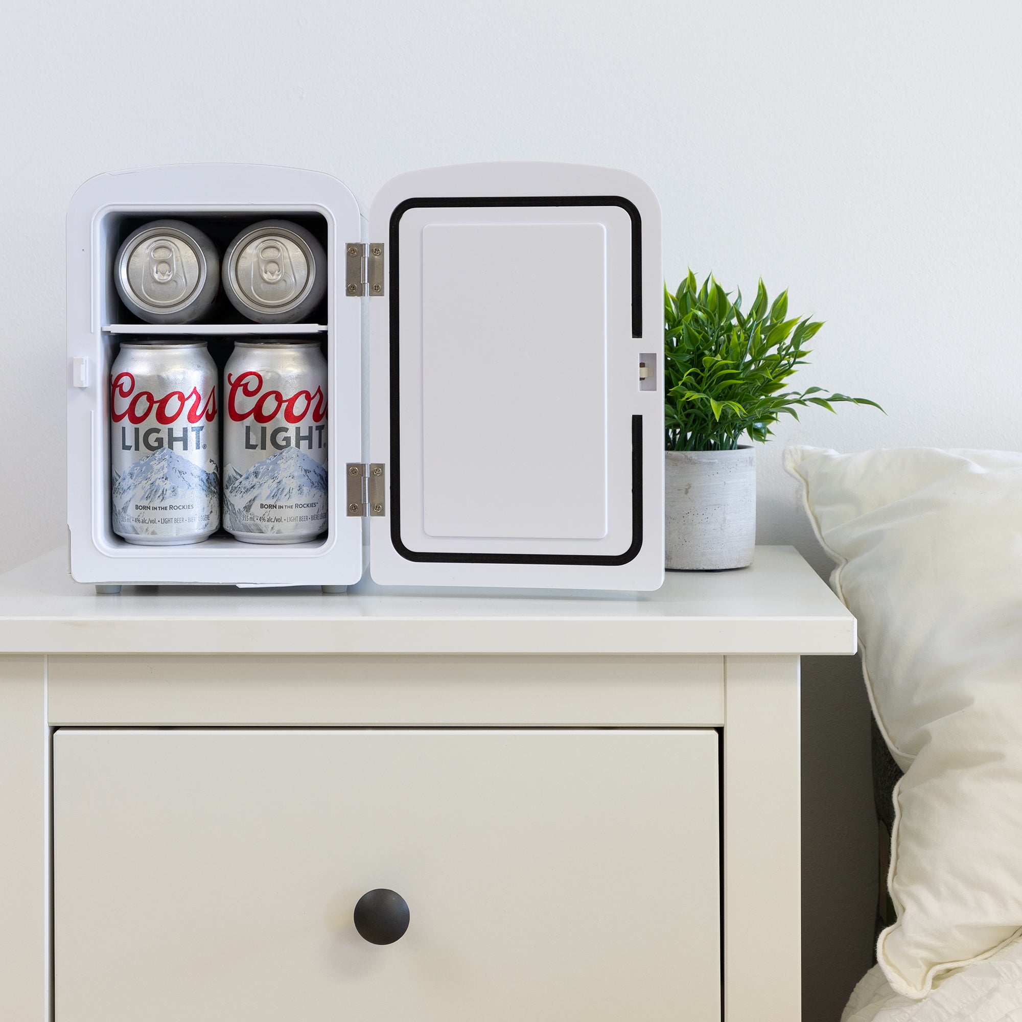 Lifestyle image of Coors Light portable mini fridge, open with 6 cans of Coors Light inside, on a white bedside table with a plant in a white pot on its right and a white painted wall behind