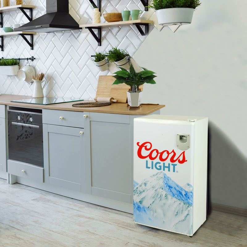 Lifestyle image of Coors Light compact fridge to the right of a gray kitchen cabinet with wooden countertop. There are potted plants, shelves, cooking utensils, and a white tile backsplash visible