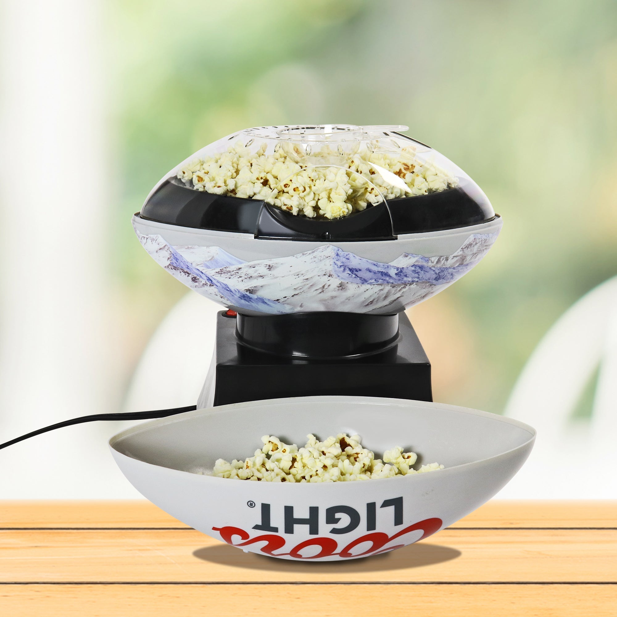 Lifestyle image of Coors Light air popper on a wooden table with popcorn popping inside and the serving bowl half full of popcorn in front