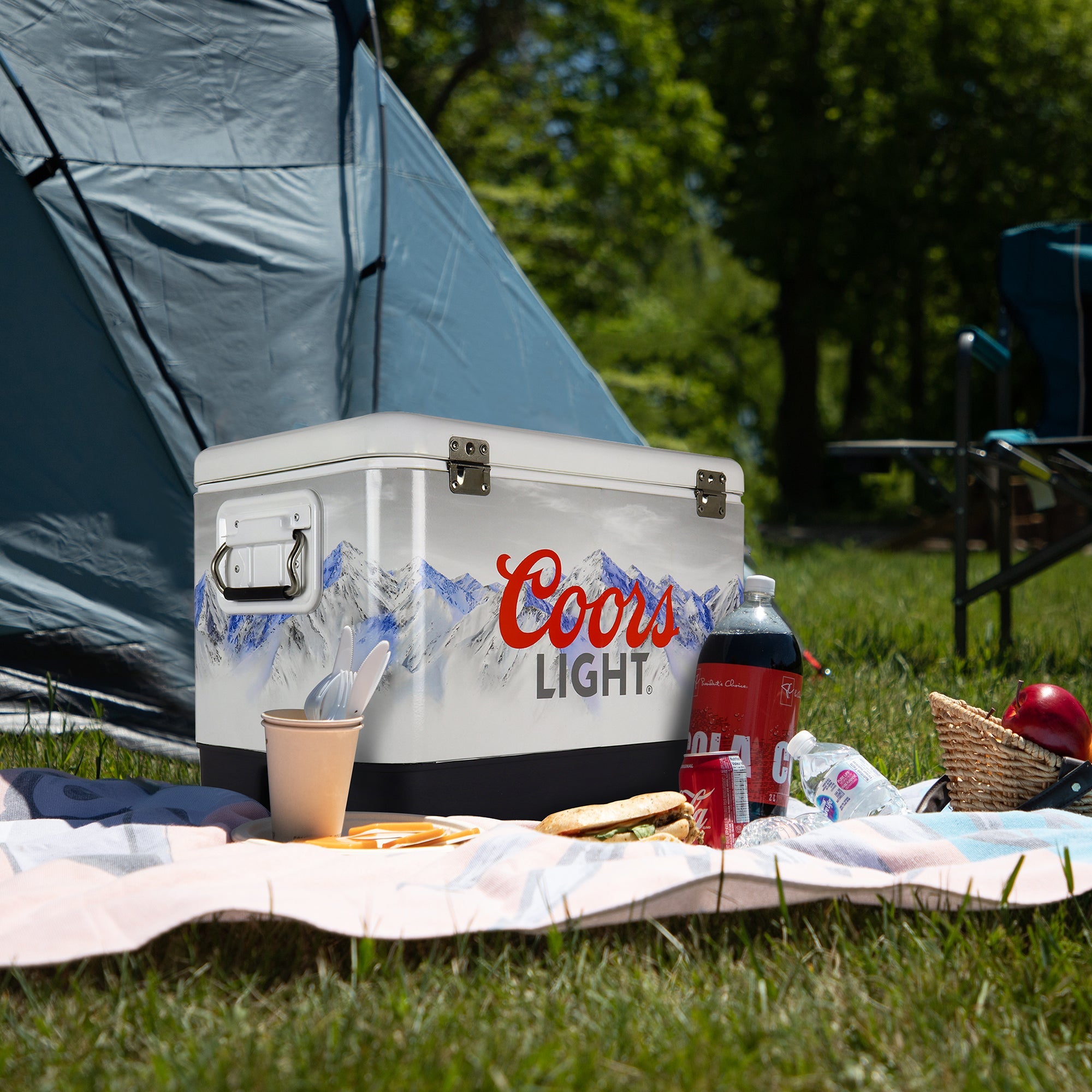 Lifestyle image of Coors Light 54 qt ice chest on a picnic blanket on the grass with fresh fruit and a bottle and can of soda in front and a teal dome tent behind