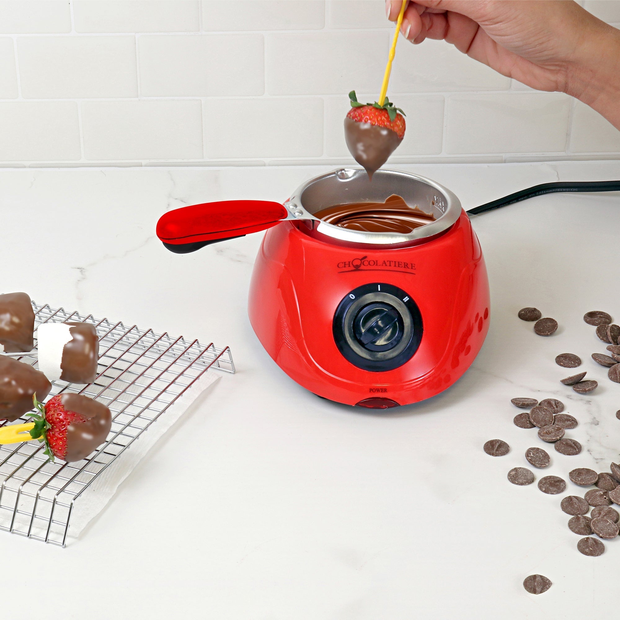 Lifestyle image of a hand dipping a strawberry in melted chocolate in the chocolatiere on a white countertop. There is a cooling rack to the left with dipped strawberries and marshmallows on it and unmelted chocolate discs scattered on the counter to the right.