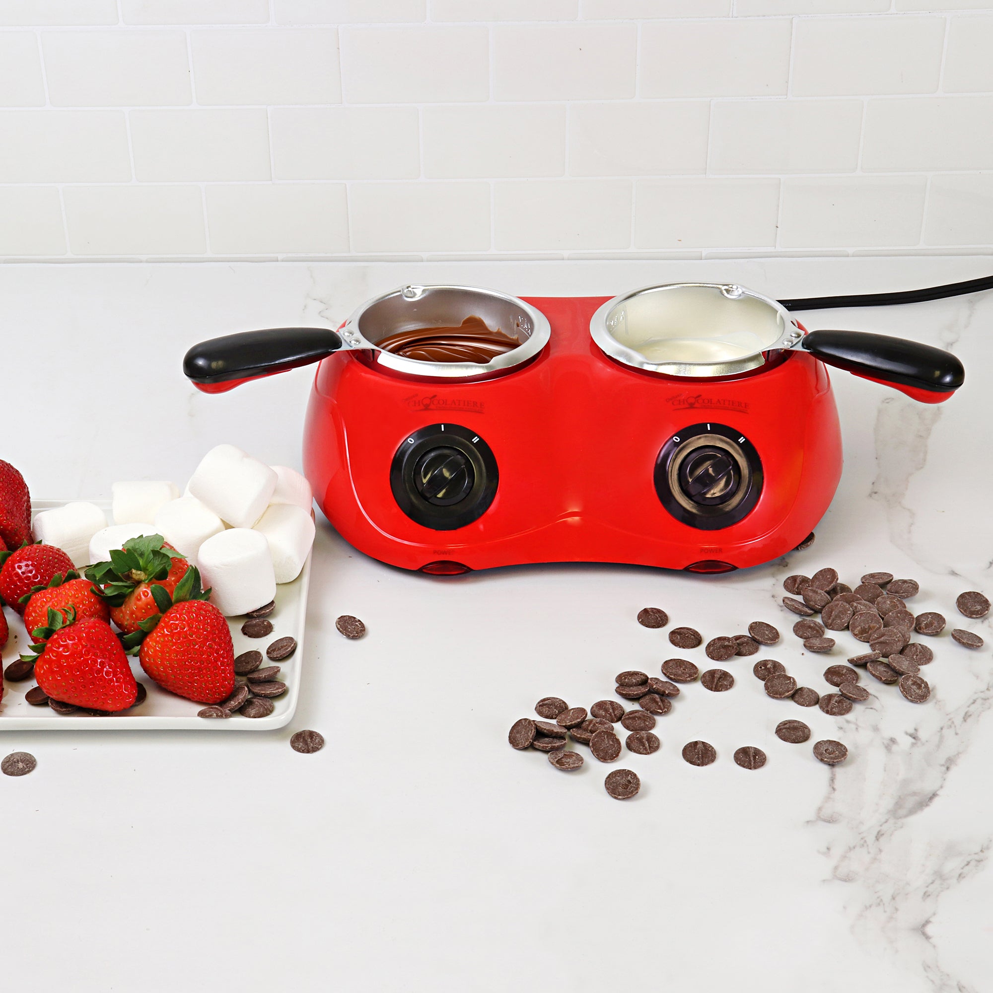 Lifestyle image of the chocolatiere on a white marbled countertop with melted milk chocolate in one pot and white chocolate in the other. There is a plate to the left with strawberries and marshmallows on it and unmelted chocolate discs scattered on the counter to the right