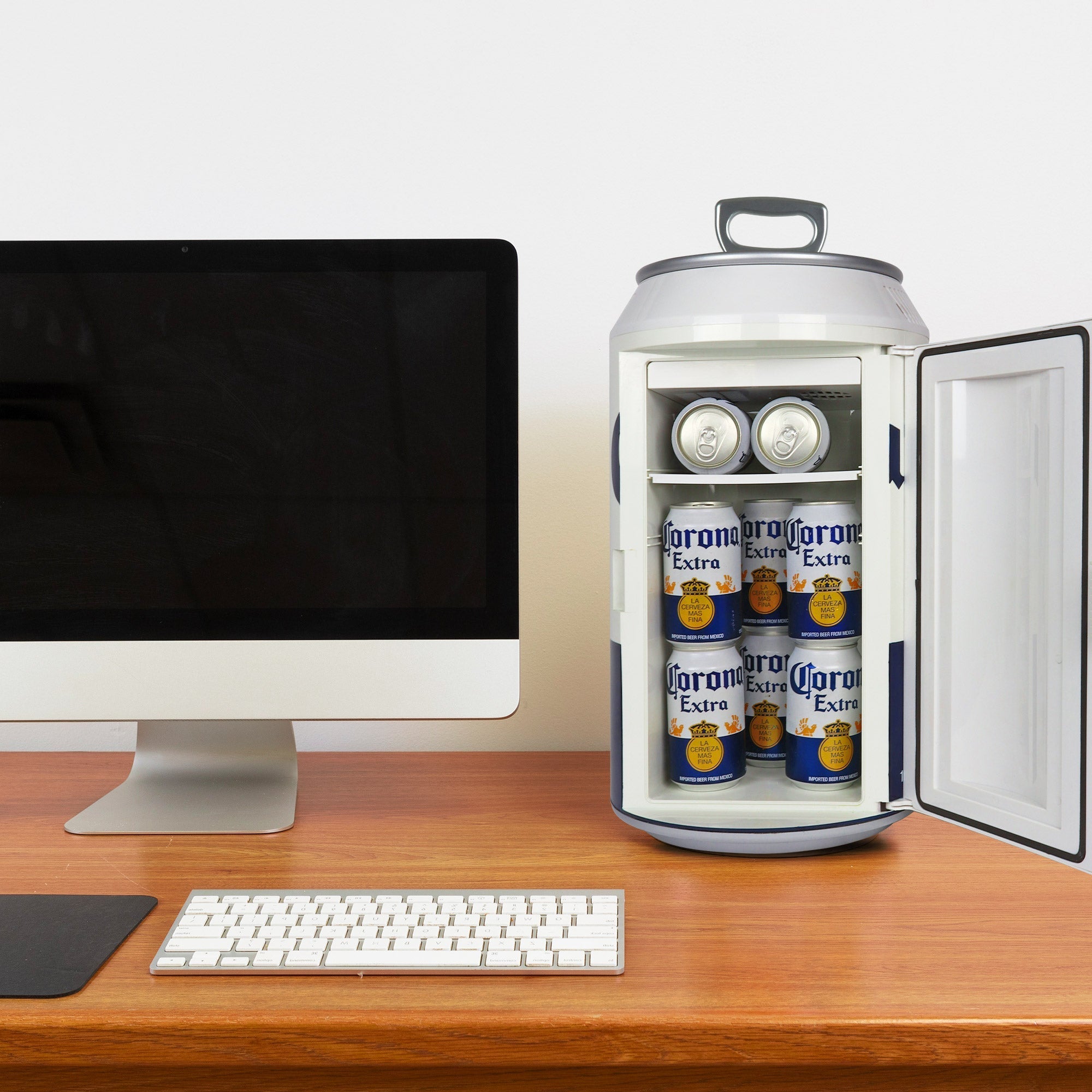 Lifestyle image of Corona can-shaped mini fridge, open with 8 cans of Corona beer inside, on a wooden desktop with a computer monitor and keyboard beside and a white wall behind
