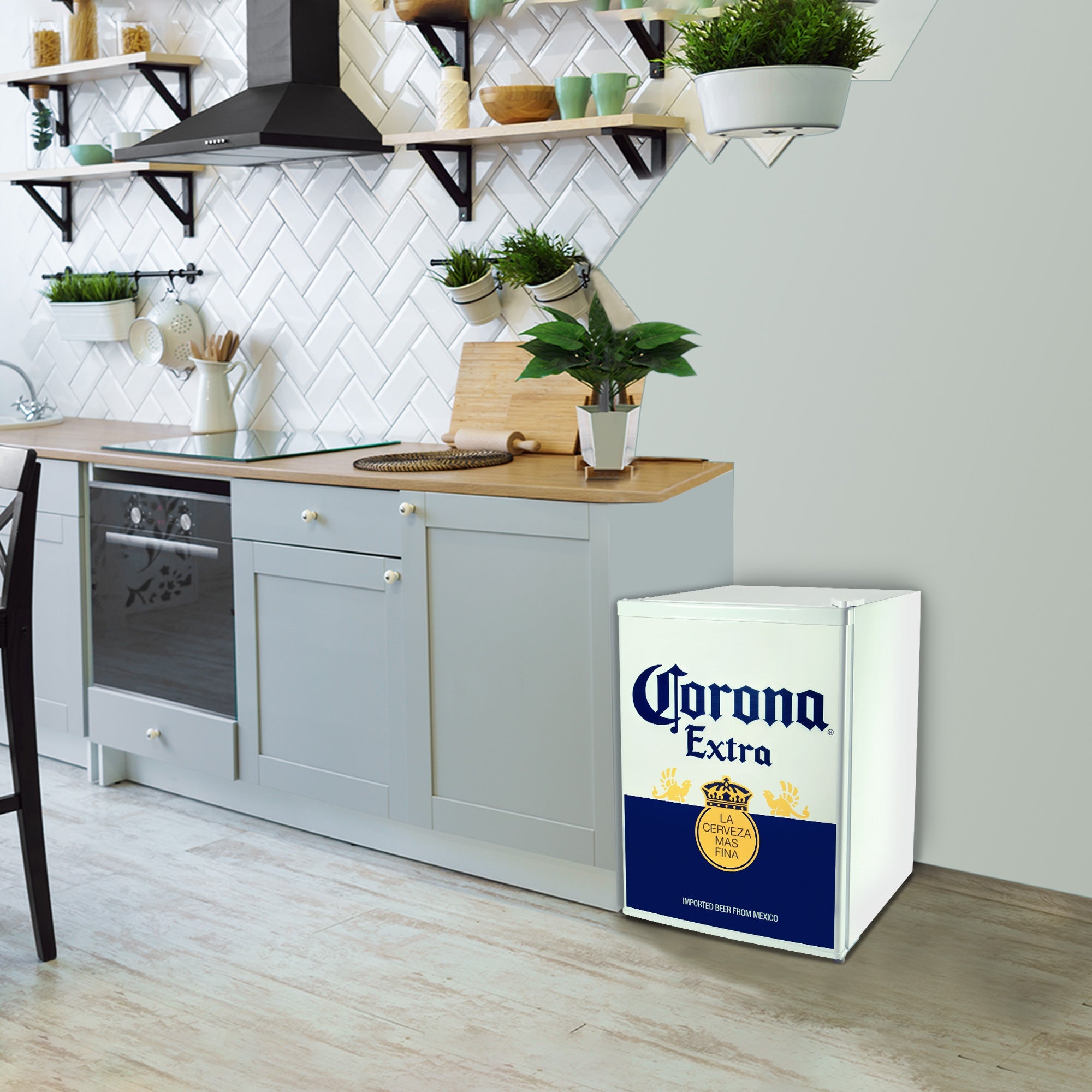 Lifestyle image of Corona compact beer fridge with freezer to the right of a set of gray cupboards with a light-coloured wooden countertop. There is a light gray wall behind with white angled tile backsplash and many potted plants in ceramic planters on the counter and shelves