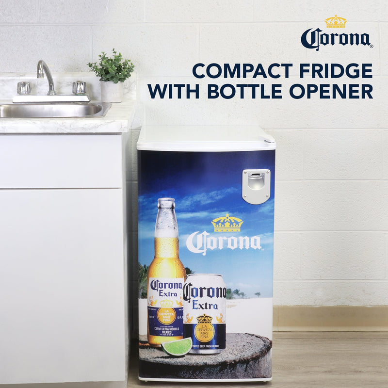 Lifestyle image of Corona compact fridge to the right of a white kitchen cabinet with white and gray marble countertop, stainless steel sink, and potted plant. Text overlay reads, "Corona compact fridge with bottle opener"