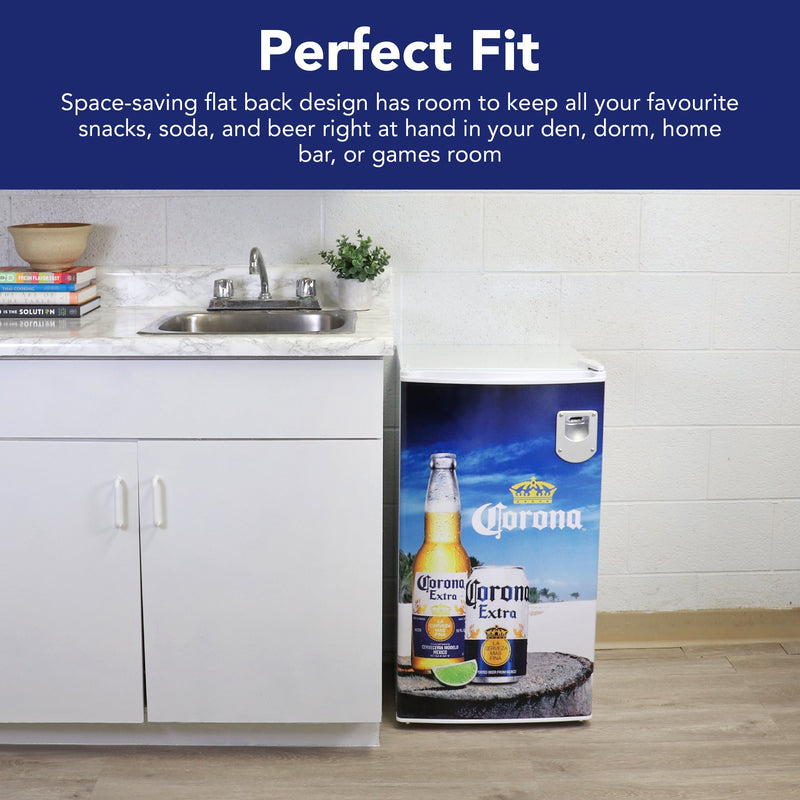 Lifestyle image of Corona compact fridge to the right of a white kitchen cabinet with white and gray marble countertop and stainless steel sink. There is a potted plant, a stack of recipe books, and a light brown ceramic bowl on the counter. Text above reads, "Perfect fit: Space-saving flat back design has room to keep all your favorite snacks, soda, and beer right at hand in your den, dorm, home bar, or games room"