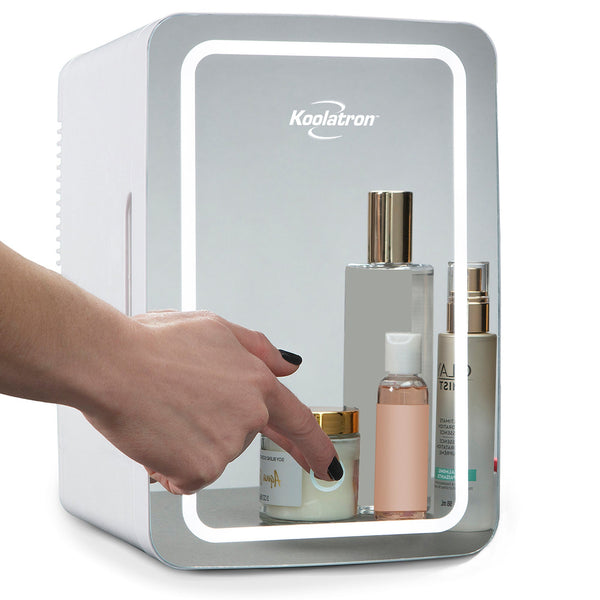 Product shot of mirrored cosmetics fridge on a white background with skincare items reflected, LED ring light illuminated, and a person's hand is visible with their index finger touching the dimmer control