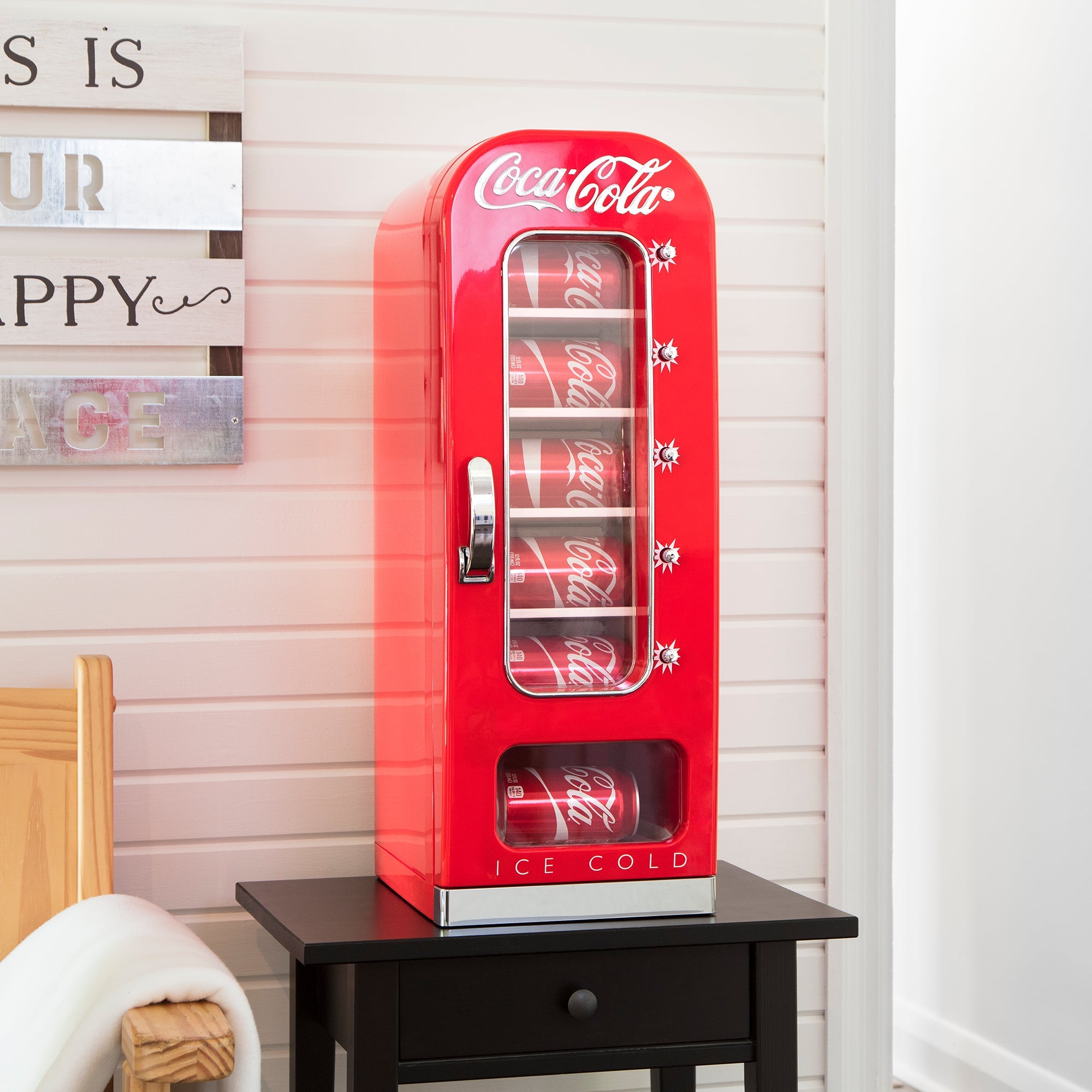 Lifestyle image of Coca-Cola 10 can vending mini fridge filled with cans of Coke on a black side table with a white shiplap wall behind and a light-coloured wooden seat to the left