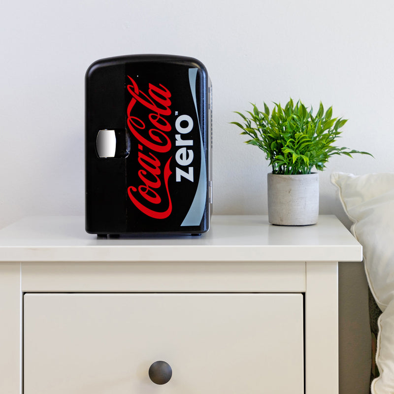 Lifestyle image of Coca-Cola Coke Zero 6 can mini fridge, closed, on a white bedside table with a plant in a white pot on its right and a white painted wall behind