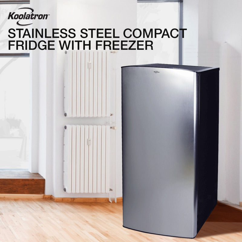 Lifestyle image of black and stainless steel compact fridge with freezer on a light-colored wood floor with a white painted wall and radiator behind. Text above reads "Koolatron stainless steel compact fridge with freezer"
