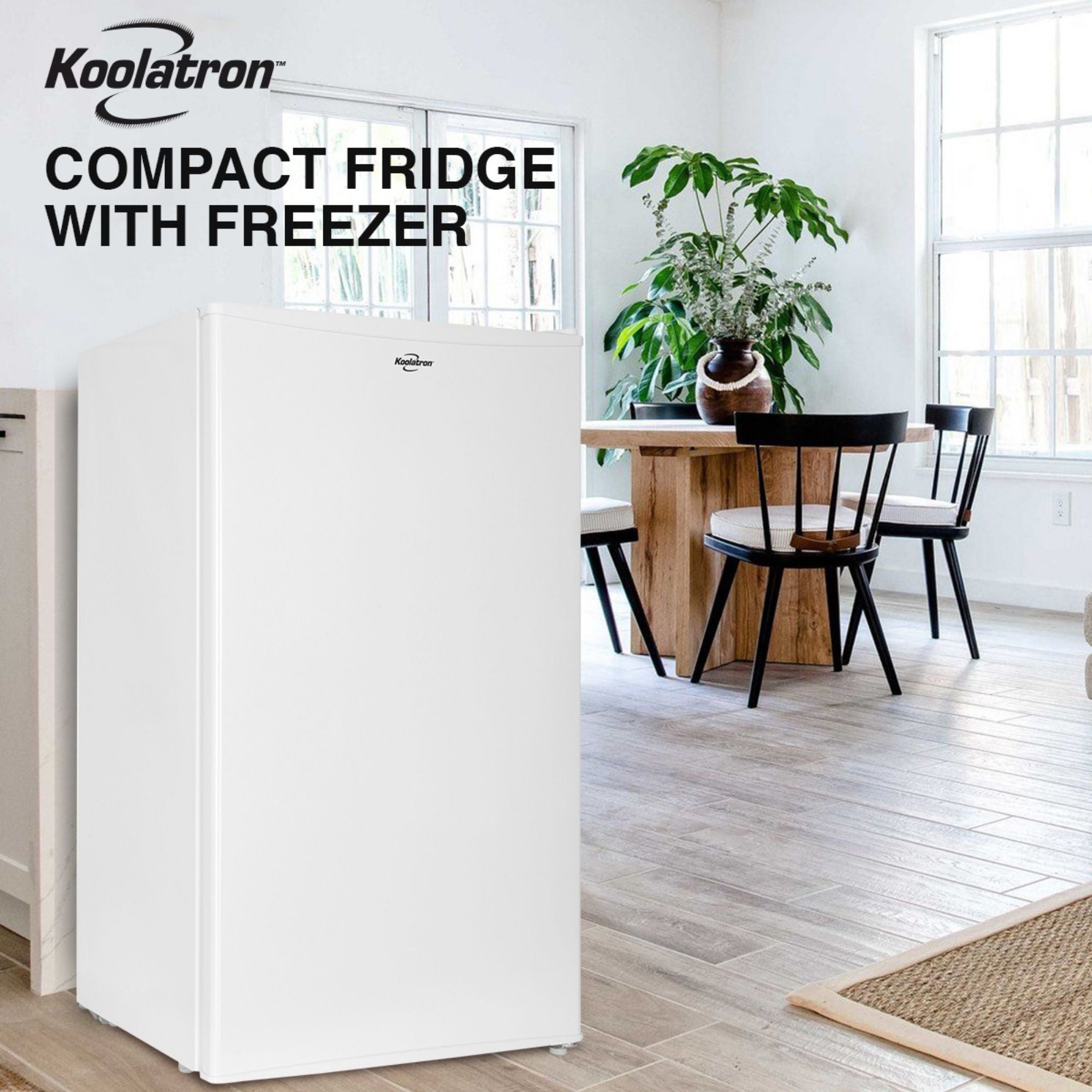 Lifestyle image of white compact fridge with freezer on a light-colored plank floor with a wooden table and chairs, two large windows, and a large potted plant in the background. Text above reads "Koolatron compact fridge with freezer"