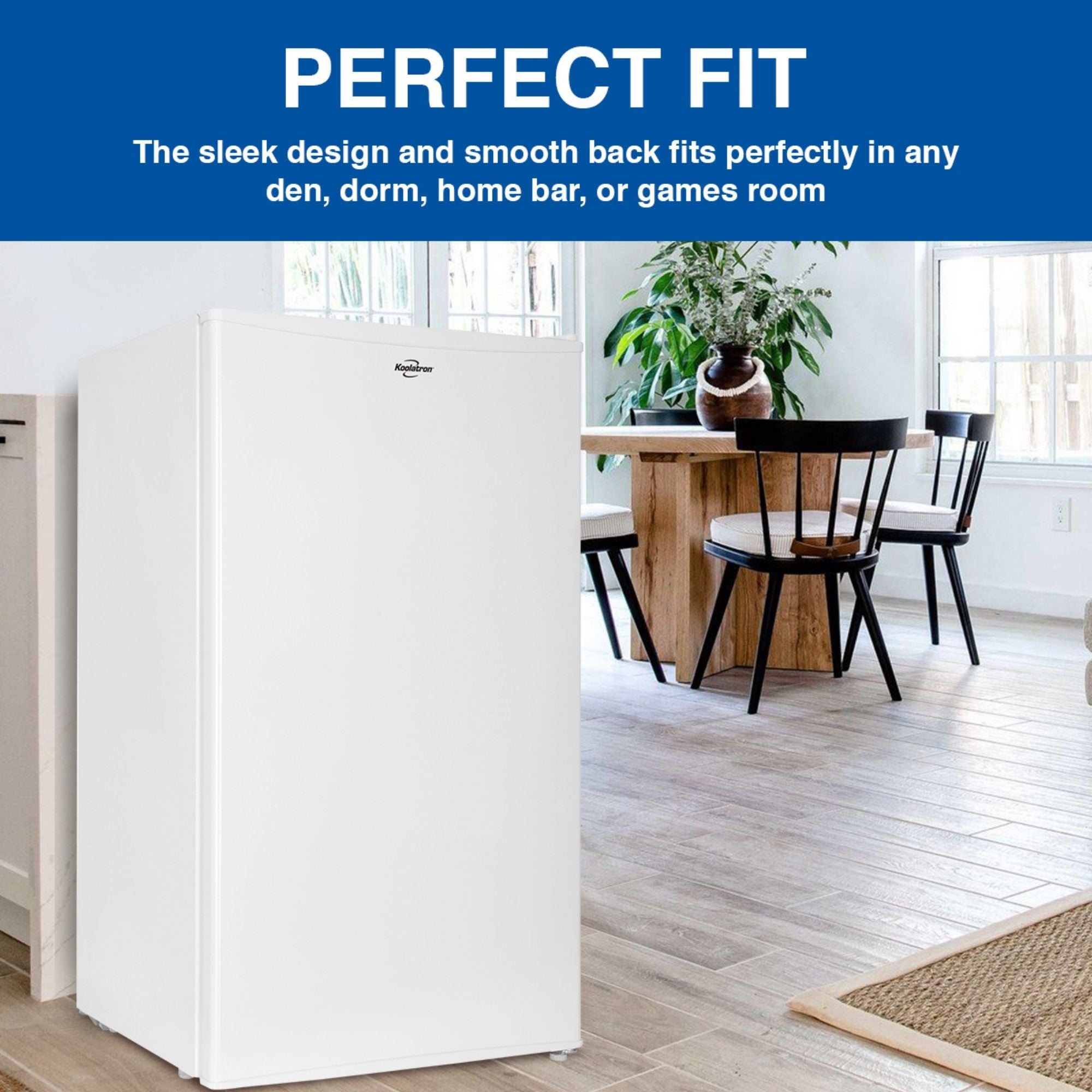 Lifestyle image of white compact fridge with freezer on a light-colored plank floor with a wooden table and chairs, two large windows, and a large potted plant in the background. Text above reads, "Perfect fit: The sleek design and smooth back fits perfectly in any den, dorm, home bar, or games room"