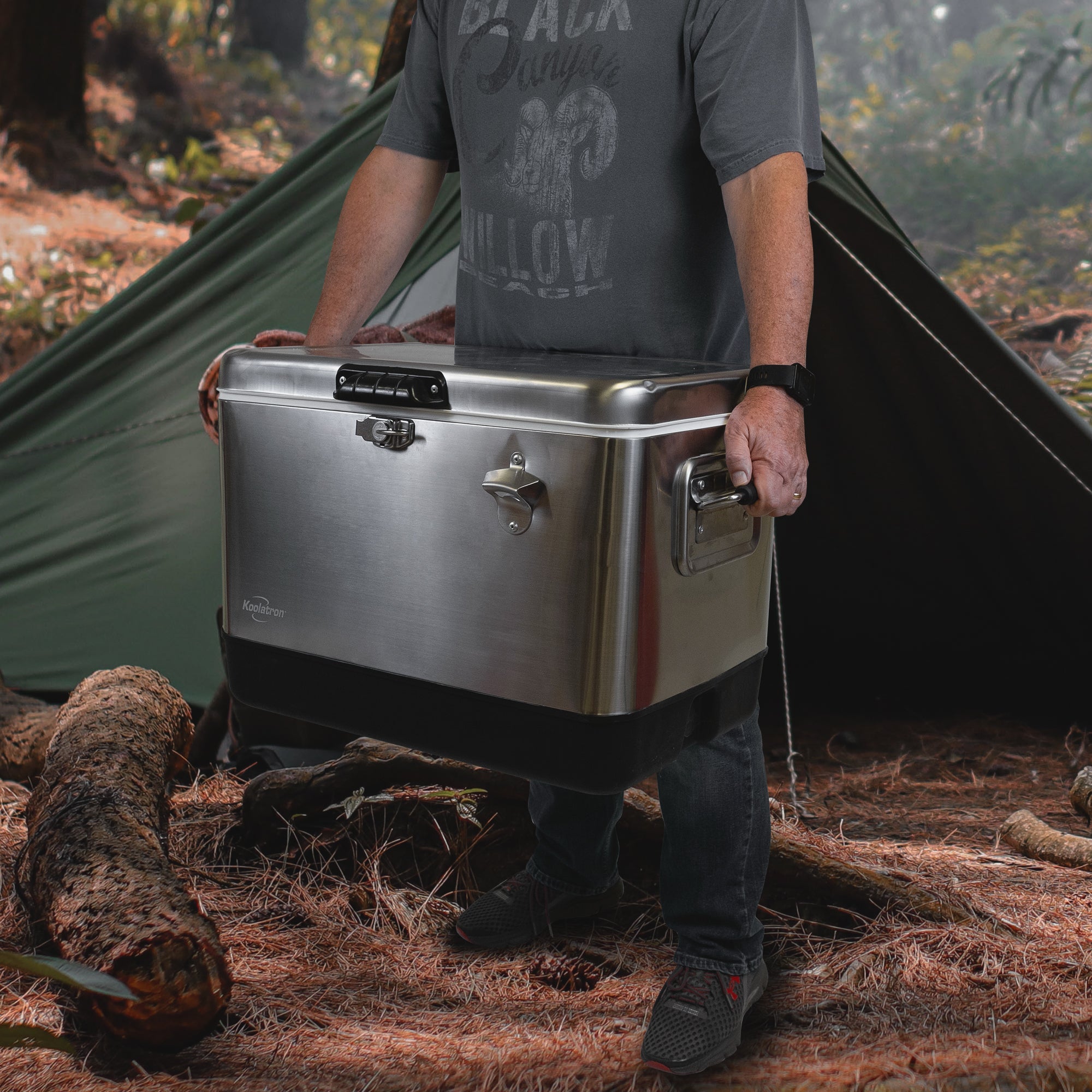 Lifestyle image of a person wearing jeans and a blue t-shirt standing up holding the Koolatron stainless steel 54 quart ice chest in front of a dark green A-frame tent