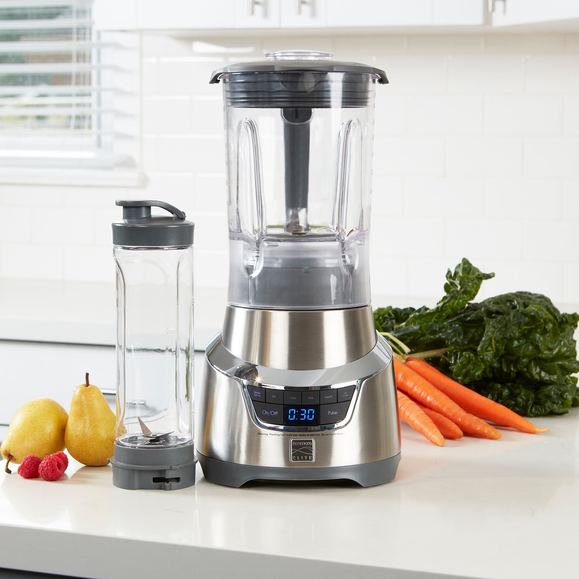 Lifestyle image of Elite 1.3 horsepower blender and travel blending cup empty on white counter with fruits and vegetables beside
