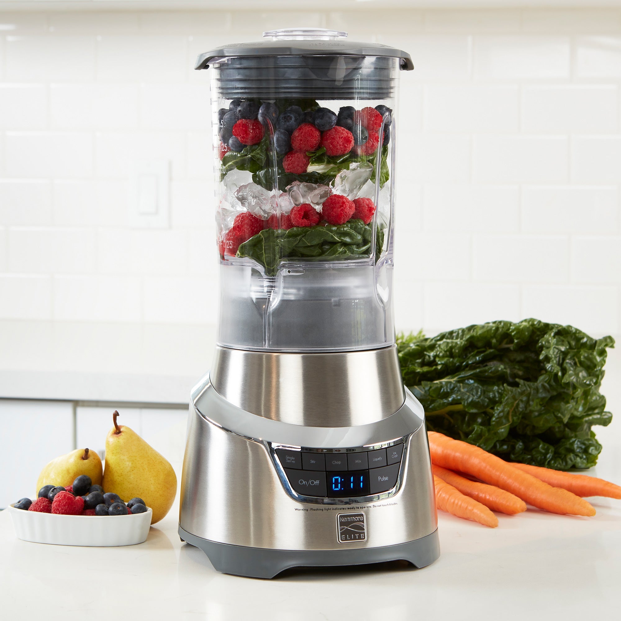 Lifestyle image of blender on white counter with layers of ice and berries ready to blend in the pitcher and fruits and vegetables beside