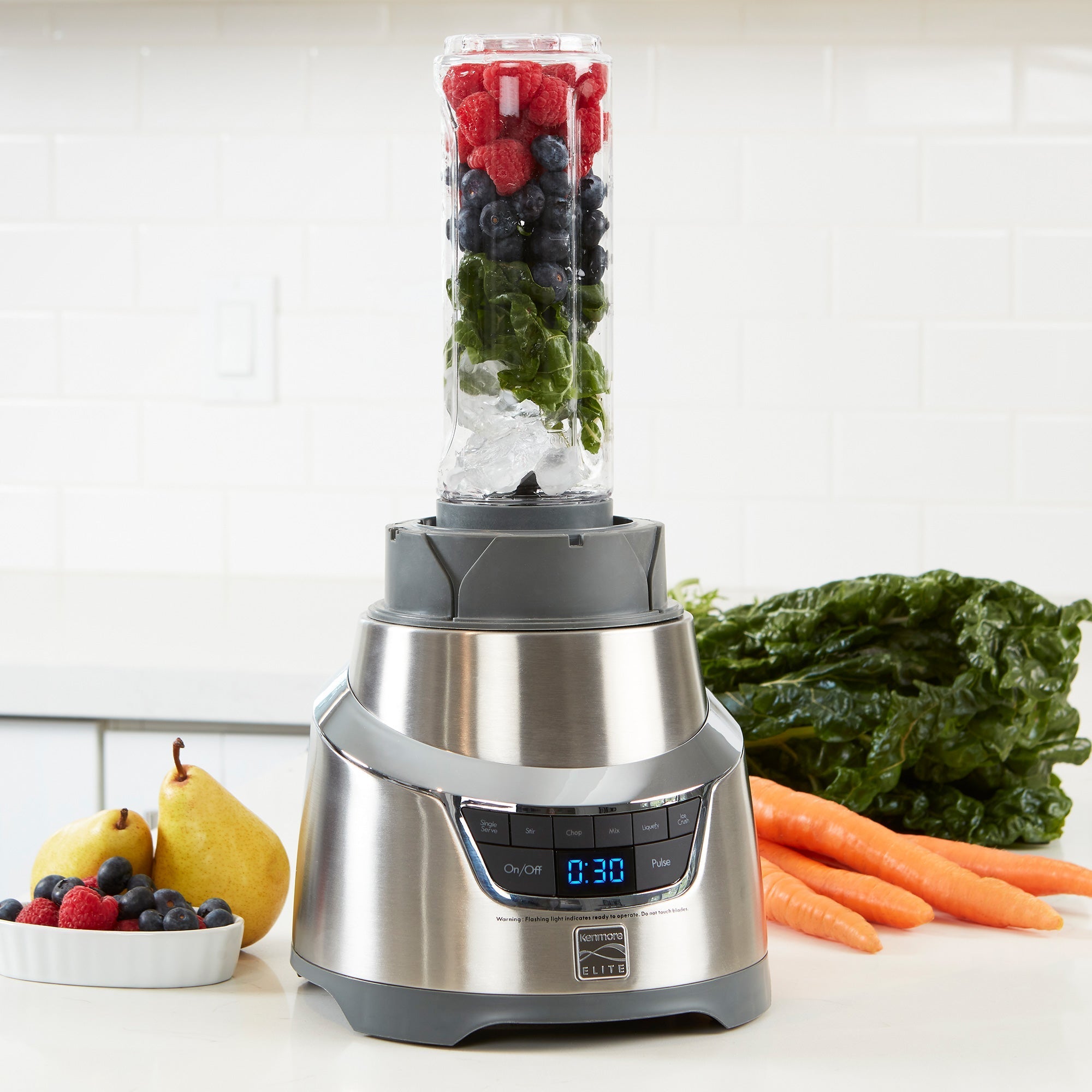 Lifestyle image of 1.3 horsepower blender on white counter with layers of ice and berries ready to blend in the single-serving cup and fruits and vegetables beside