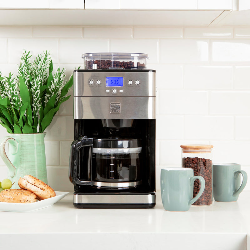 Lifestyle image of Kenmore grind and brew coffee maker filled with brewed coffee on a white quartz countertop with white tile backsplash. There is greenery in a ceramic pitcher and a plate with sliced bagels to the left and a glass jar of coffee beans and two light blue mugs to the right