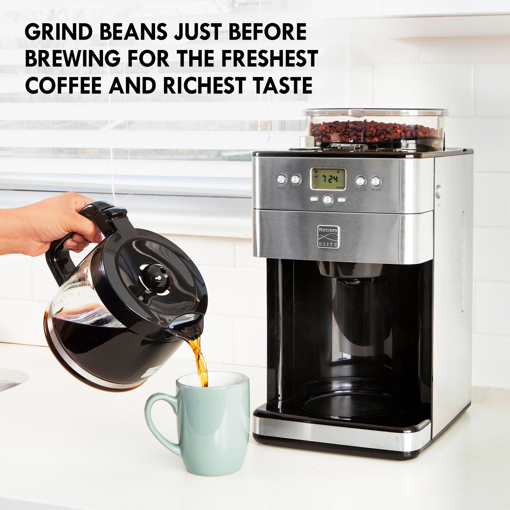 Lifestyle image of Kenmore grind and brew coffee maker on a white counter with white tile backsplash and a window with white blinds behind it and a person's hand pouring coffee from the glass carafe into a light blue mug in front. Text above reads, "Grind beans just before brewing for the freshest coffee and richest taste"