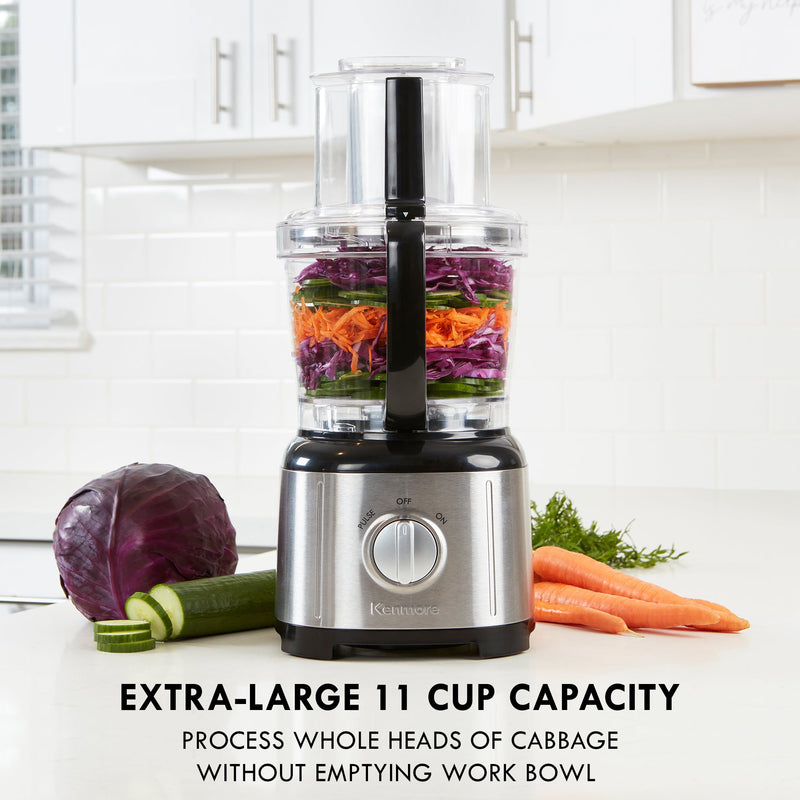 Lifestyle image of Kenmore 11 cup food processor and vegetable chopper on a light gray countertop with white tile backsplash and cupboards behind. There are chopped vegetables in the food processor and a whole red cabbage, partially sliced cucumber, carrots, and dill on either side. Text below reads, "Extra-large 11 cup capacity: Process whole heads of cabbage without emptying work bowl"