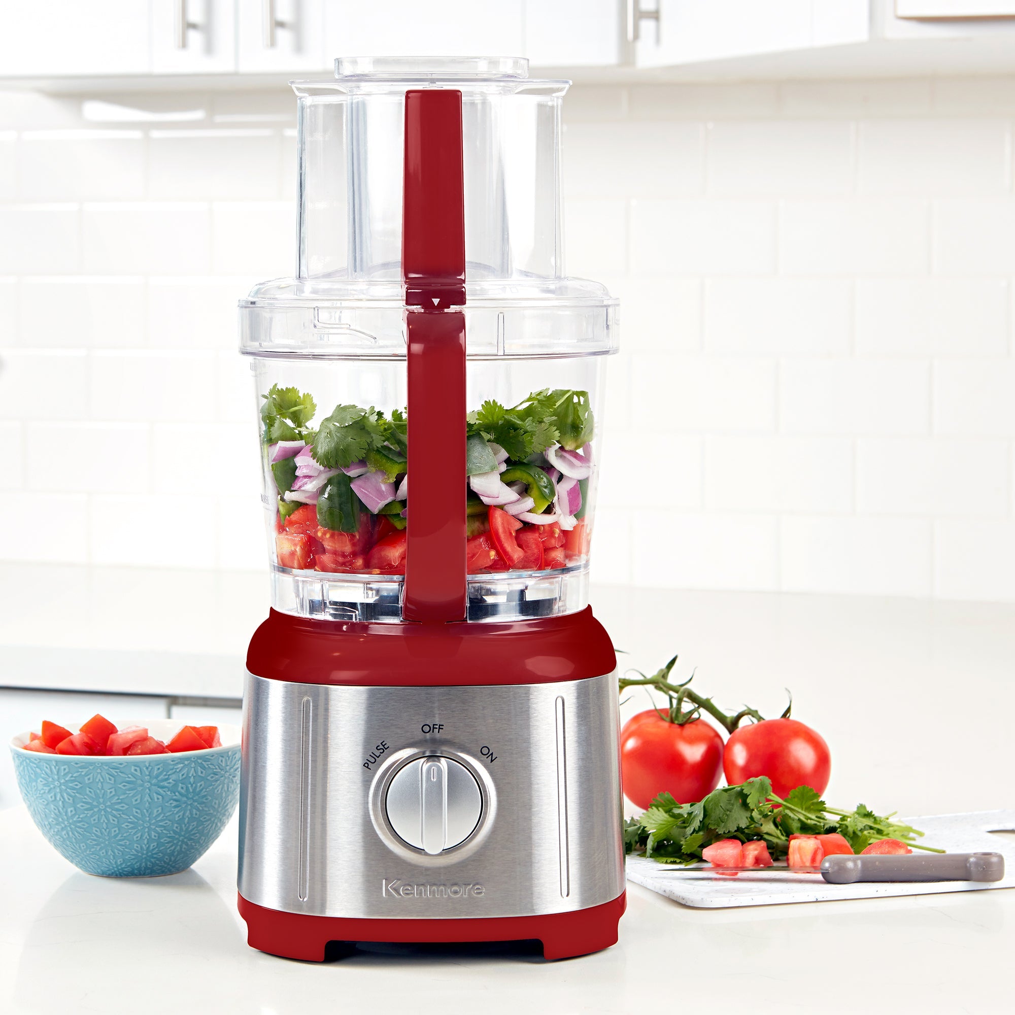 Kenmore 11 cup food processor and vegetable chopper on a light gray countertop with white tile backsplash behind. There are chopped tomatoes, red onions, and cilantro in the food processor, a bowl of chopped tomatoes to the left and a cutting board, knife, and whole and chopped ingredients to the right