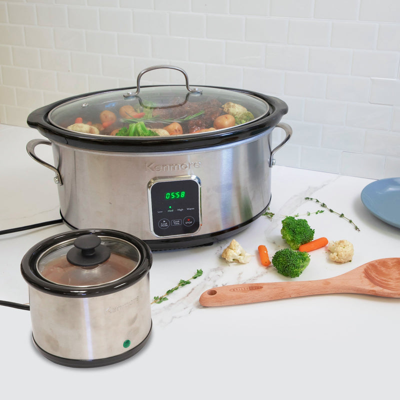 Lifestyle image of Kenmore 7 qt programmable slow cooker and sauce warmer on a white marbled countertop with white tile backsplash behind. The slow cooker contains a roast and vegetables and there are pieces of broccoli and carrots, fresh thyme sprigs, and a wooden spoon on the counter beside it