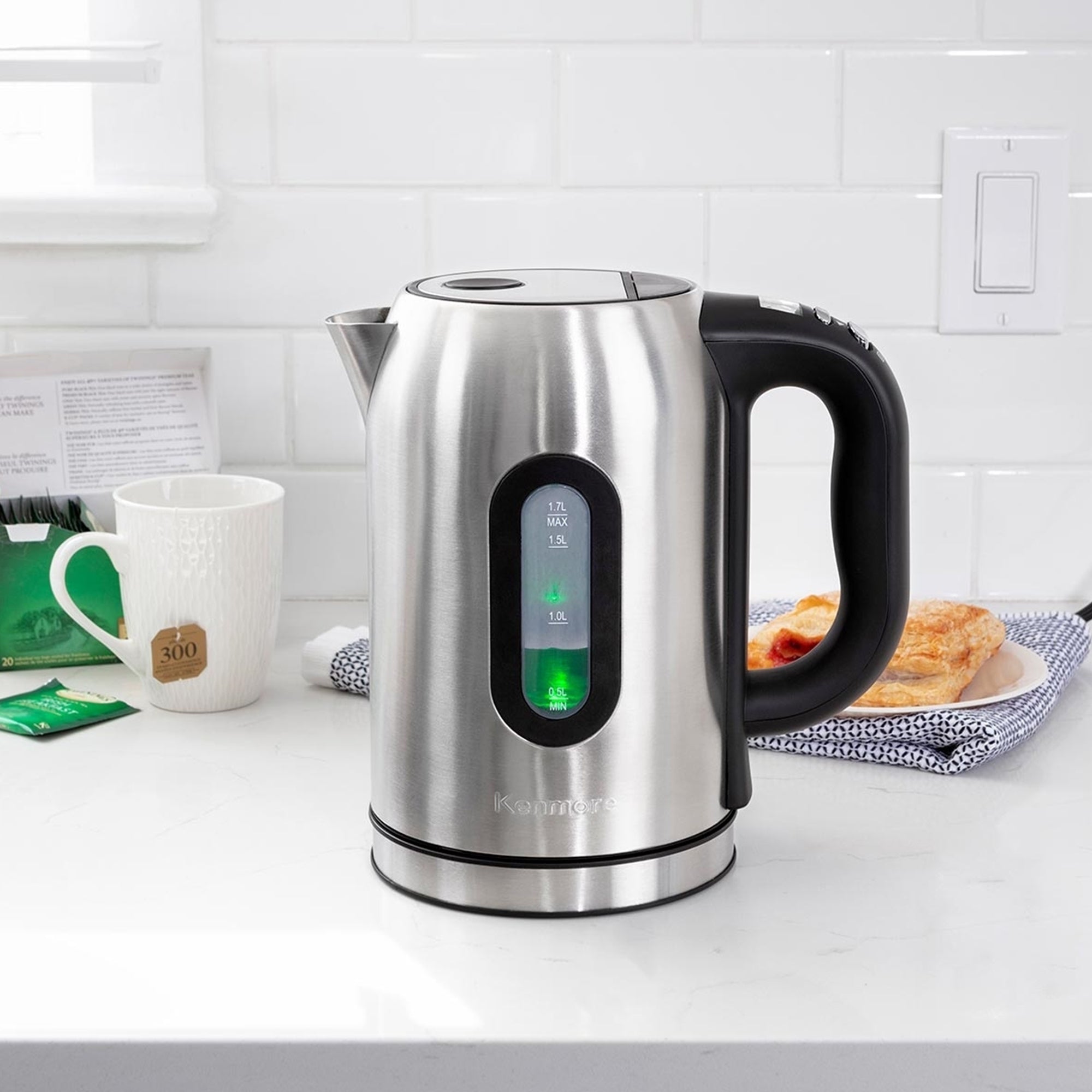 Lifestyle image of Kenmore stainless steel digital teakettle on a light gray countertop with white tile backsplash behind and a mug, teabag, pastry, and tea towel arranged around it