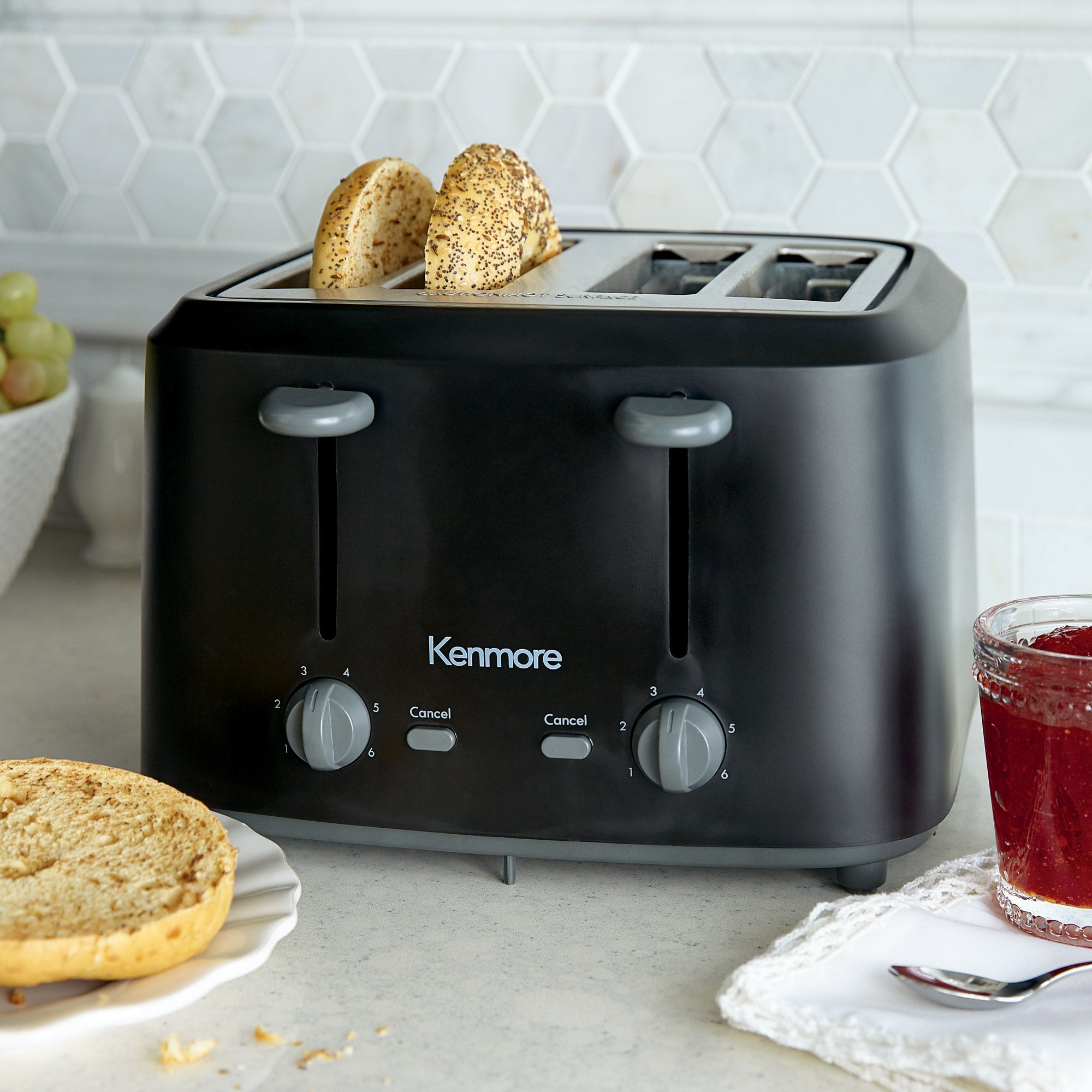 Kenmore 4-slice matte black toaster with two bagel slices inside on light gray countertop with a white marbled backsplash behind and a plate with bagel half and napkin with jam jar and spoon in front