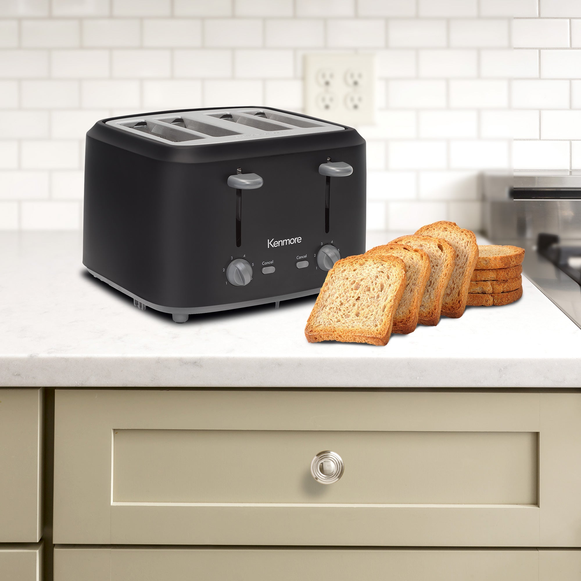 Lifestyle image of Kenmore OPP 4-slice matte black toaster on light gray countertop over a beige drawer with a white tiled backsplash behind and slices of bread in front