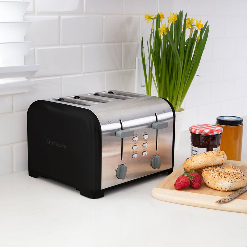 Lifestyle image of Kenmore 4-slice black stainless steel toaster on light gray countertop with white tile backsplash and a pot of yellow daffodils behind. On the right is a wooden cutting board with strawberries, uncut bagels, knife, and two jam jars