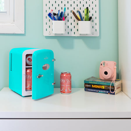 Lifestyle image of Koolatron retro 6 can mini fridge, partly open with cans inside, on a white desktop with an aqua wall behind. There is a can of soda, a stack of books, and a pink camera to the right of the fridge and two cups of pens and pencils attached to the wall above
