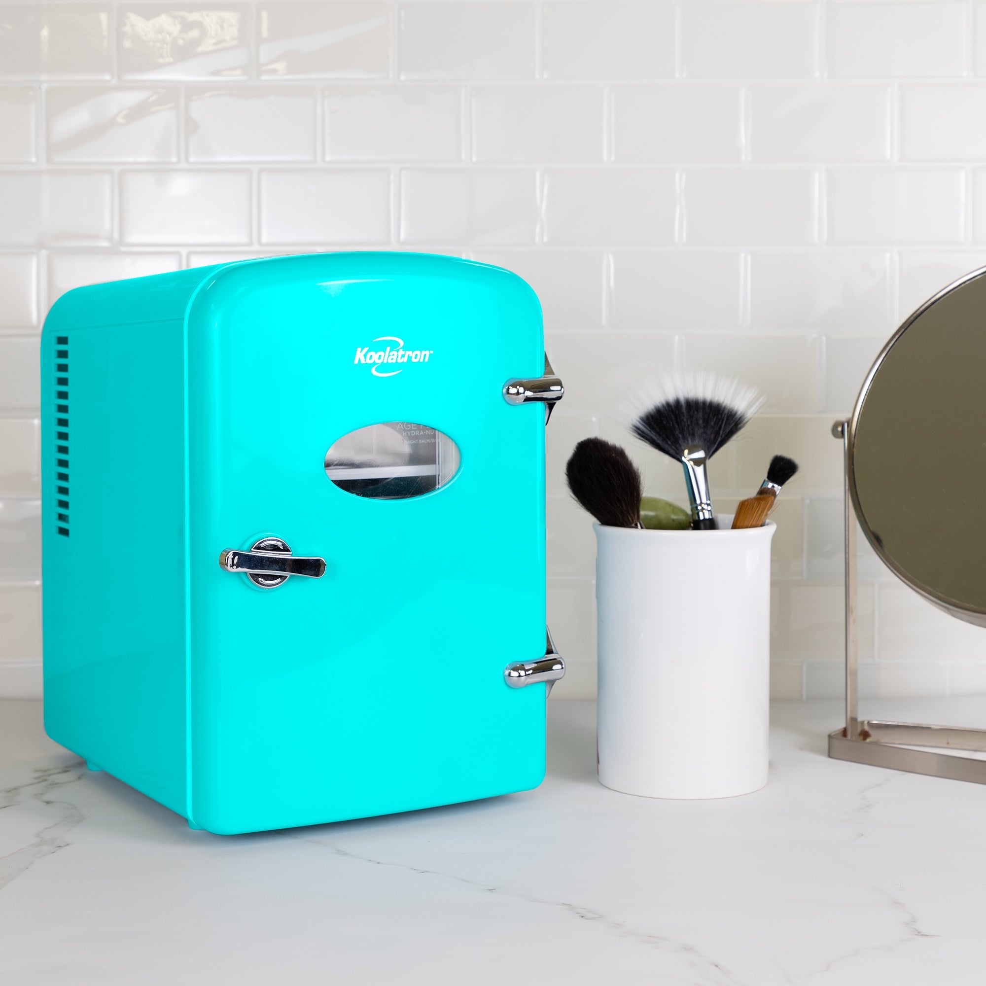 Lifestyle image of Koolatron retro 6 can mini fridge, closed, with makeup brushes in a white container on its right, on a white marbled counter with a white tile backsplash behind 