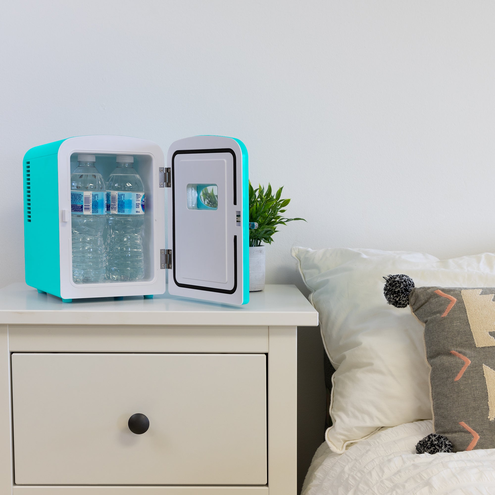 Lifestyle image of Koolatron retro 6 can mini fridge, open with 4 water bottles inside, with a plant in a white pot on its right, on a white bedside table beside a bed with white linens